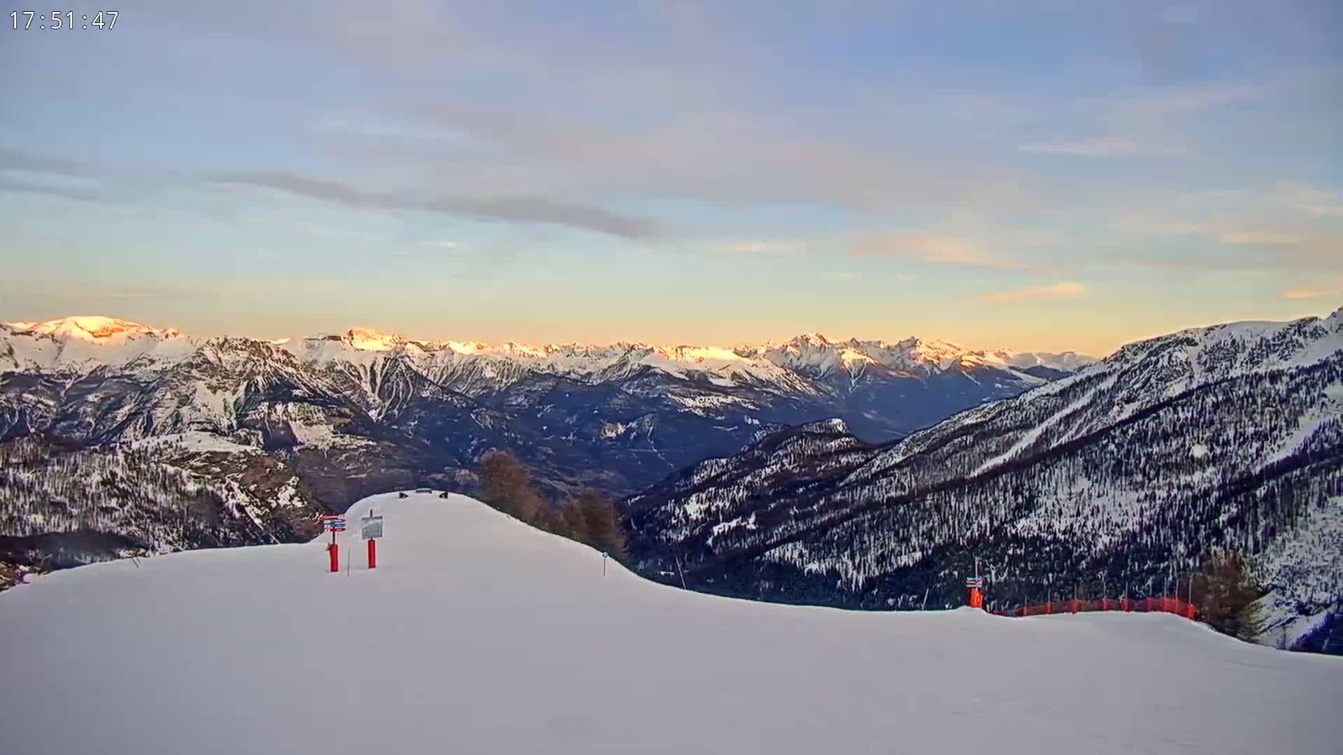 A snow-covered mountain slope overlooks a vast range of snow-capped mountains at sunset under a mostly clear sky.