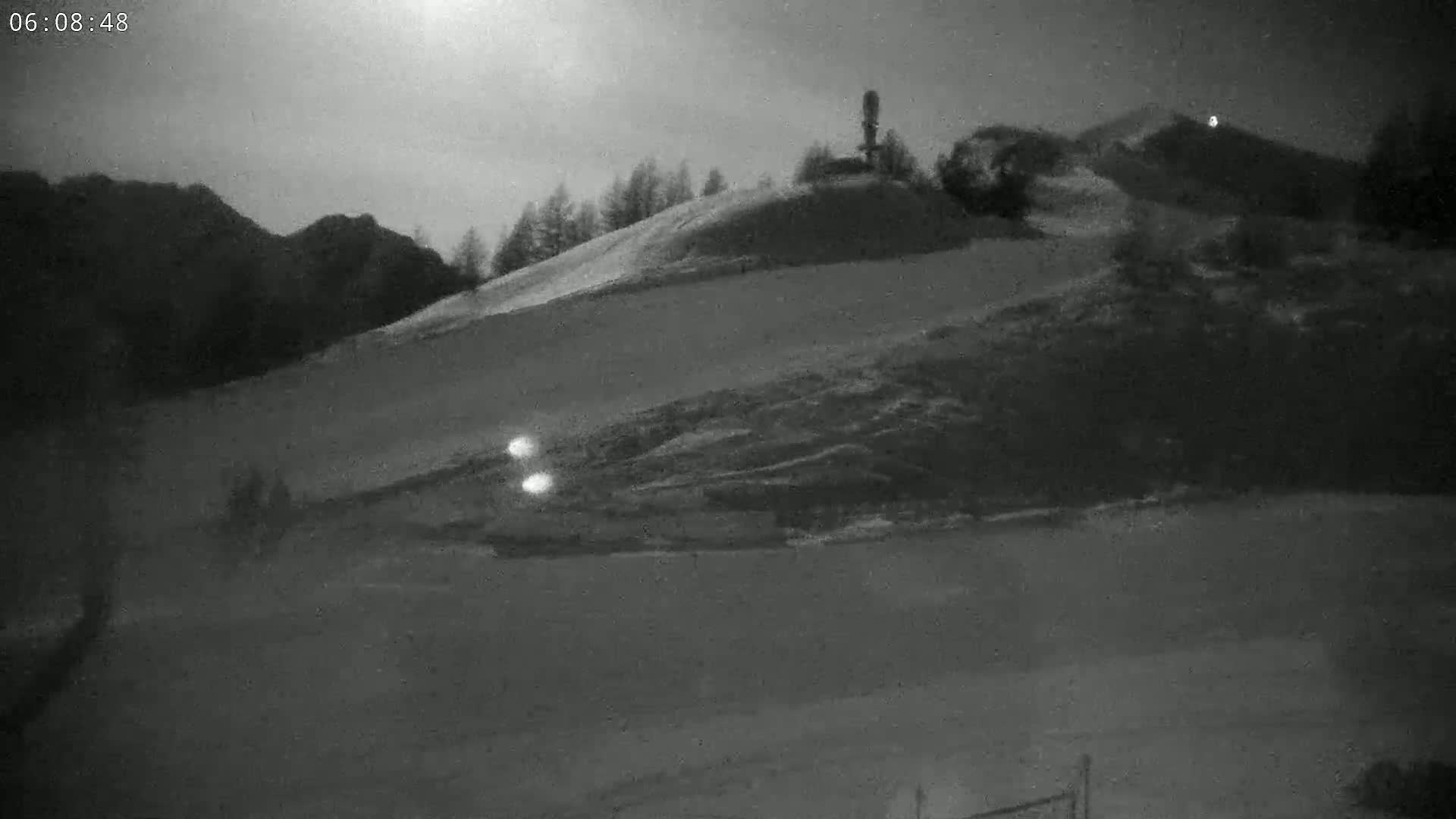 A nighttime, snow-covered hillside is illuminated by faint light, with a few small lights visible in the lower middle ground.