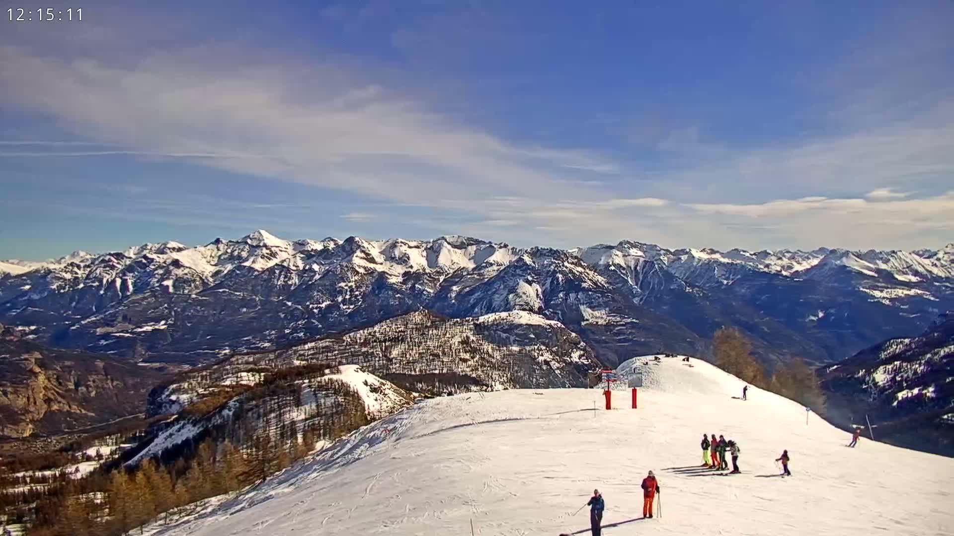 A snow-covered mountain slope with several skiers and a view of a distant, snow-capped mountain range under a mostly clear, bright sky.