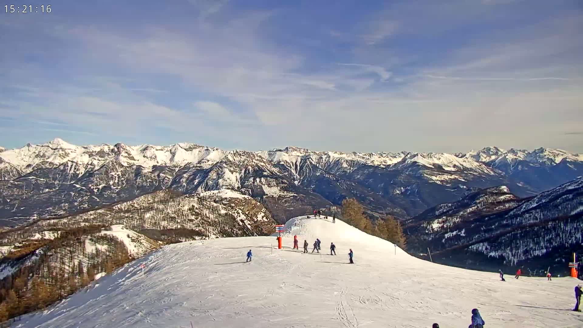 A snow-covered ski slope with several skiers and snowboarders, overlooking a vast mountain range under a mostly clear, bright sky.