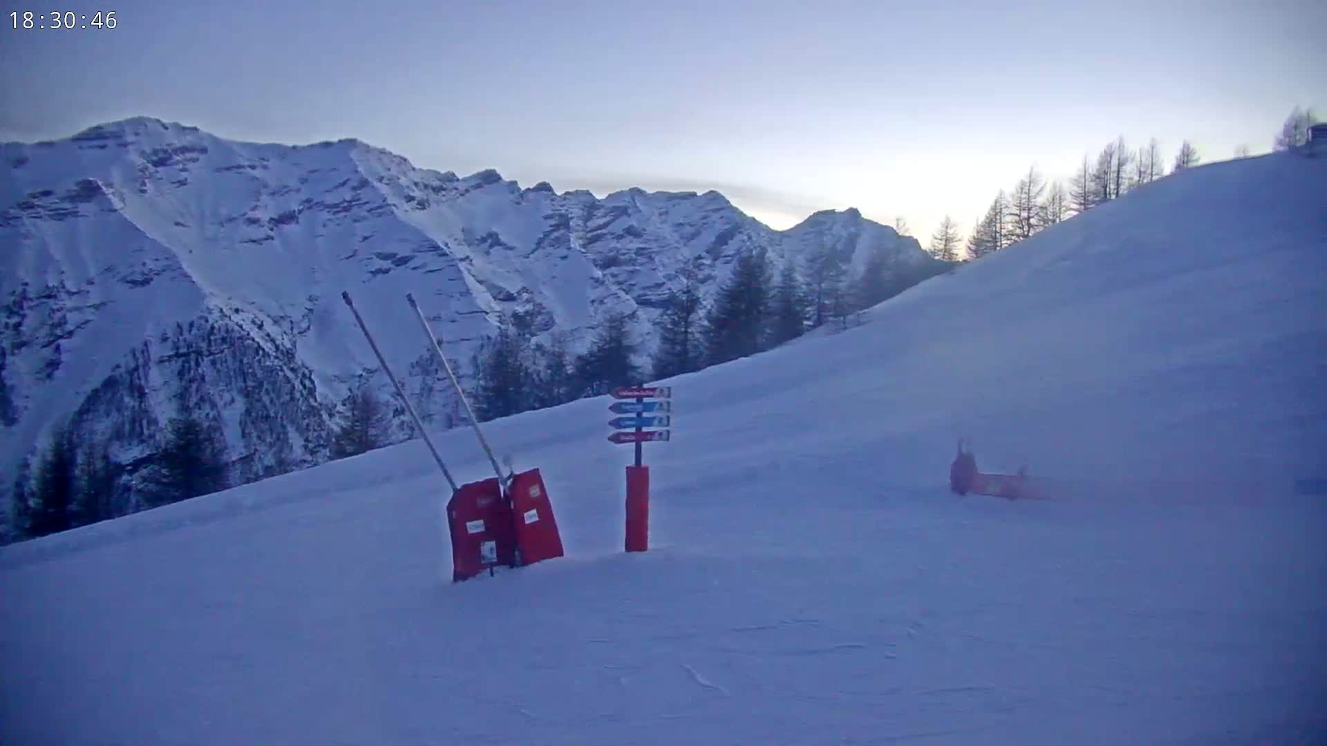 A snowy mountain slope at dusk, showing a ski run with a trail marker and some equipment.