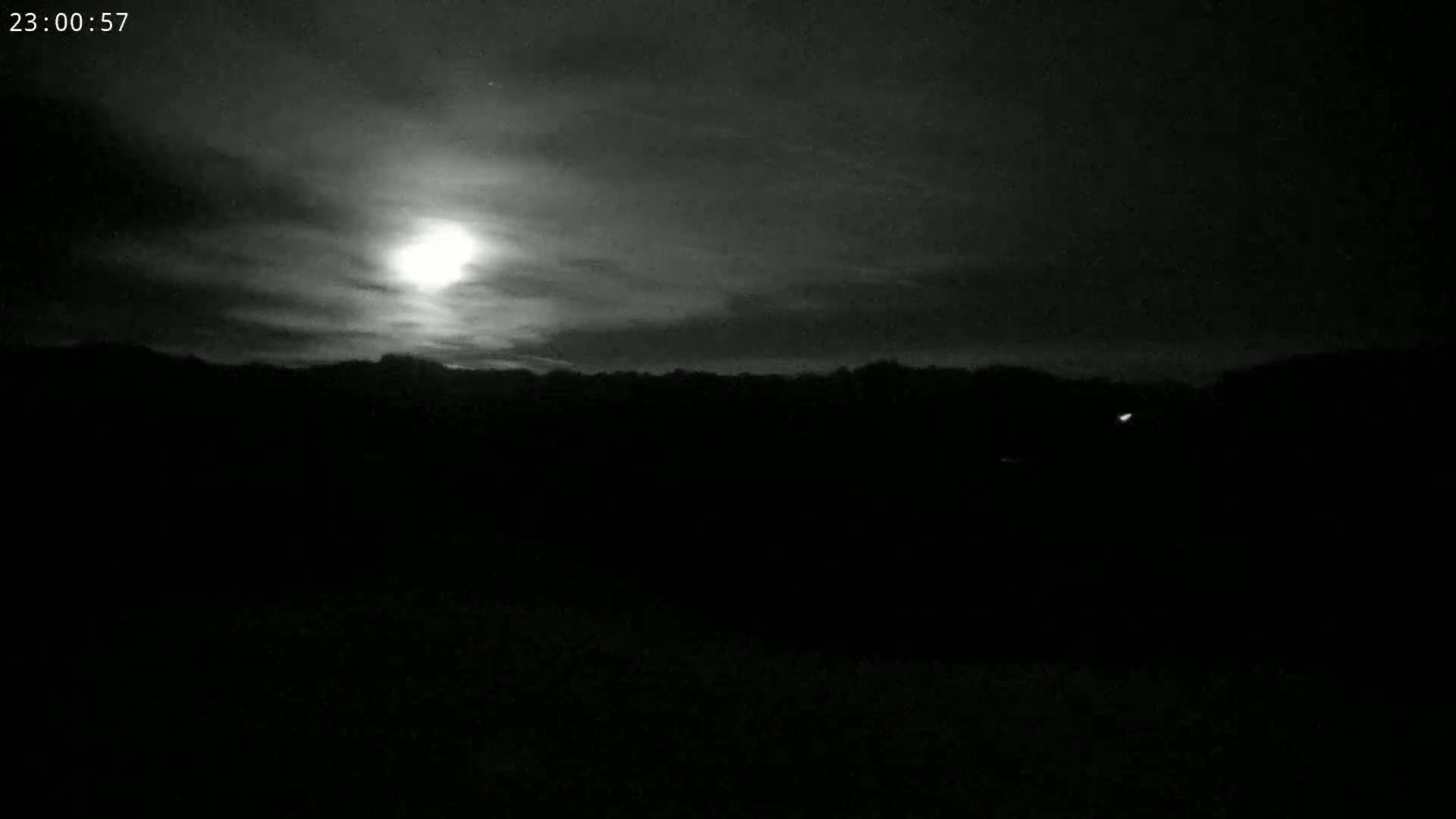A bright moon shines through a mostly cloudy night sky above a dark, silhouetted landscape.