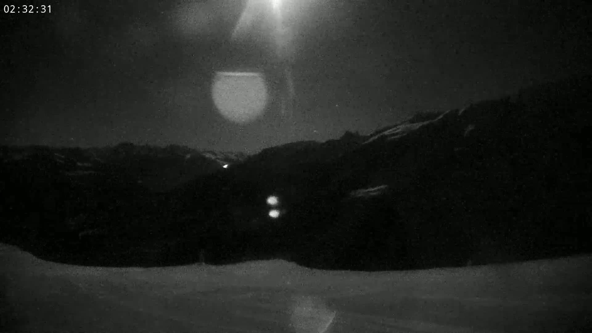 A nighttime, snowy landscape shows a dark mountain range under a bright moon or light source.