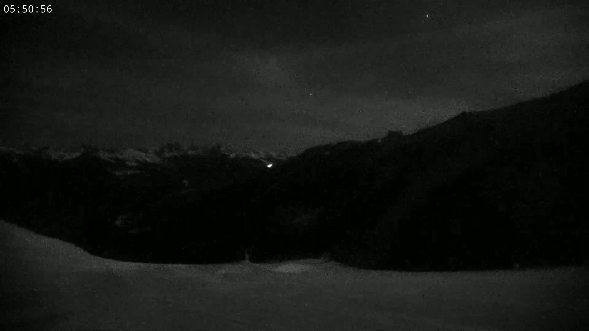A nighttime, low-light view of a snow-covered landscape with dark hills and a faint light source in the distance under a cloudy sky.