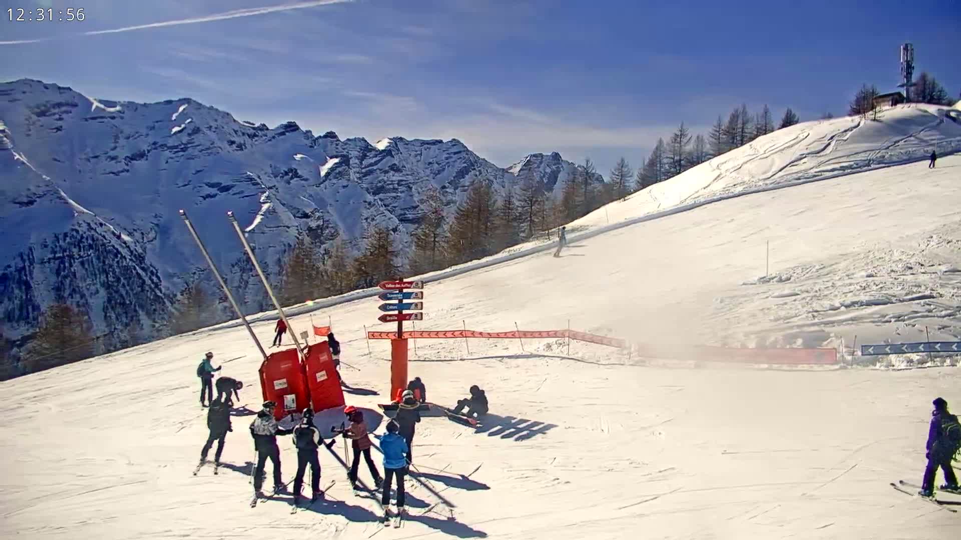 A sunny day on a snow-covered ski slope with skiers and snowboarders near a lift and signpost, set against a backdrop of snow-capped mountains.