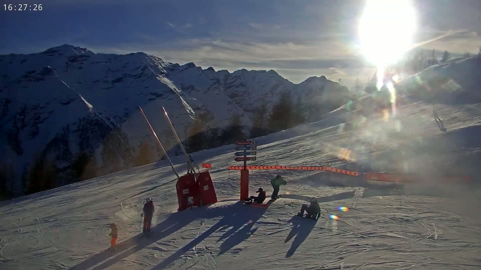 A sunny day on a snow-covered mountain slope shows several skiers and snowboarders, some resting near a signpost.