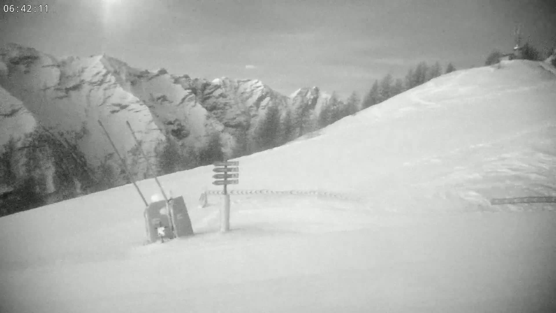 A snowy, overcast mountain slope features a trail marker and a small structure near the bottom of the hill.