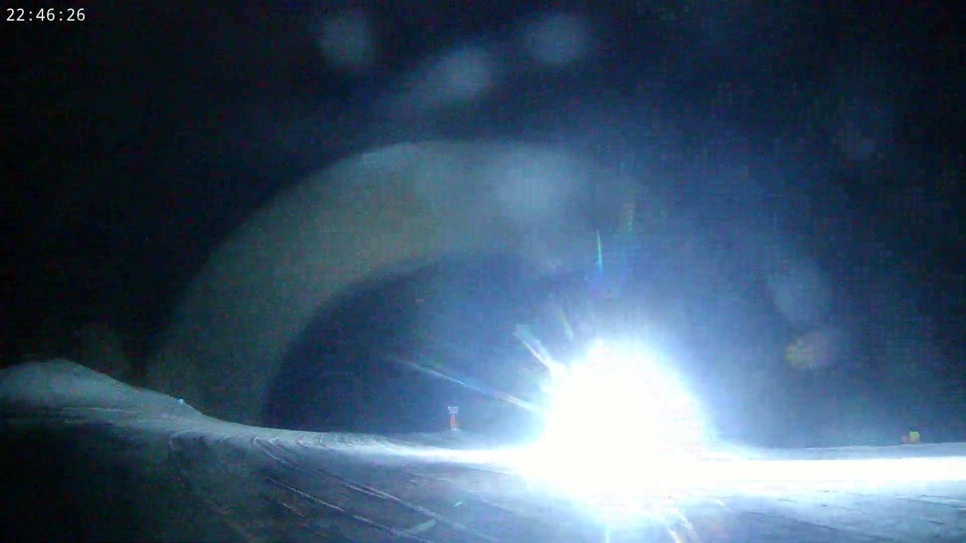 A bright light illuminates a snow-covered slope at night, with blowing snow obscuring the surrounding area.