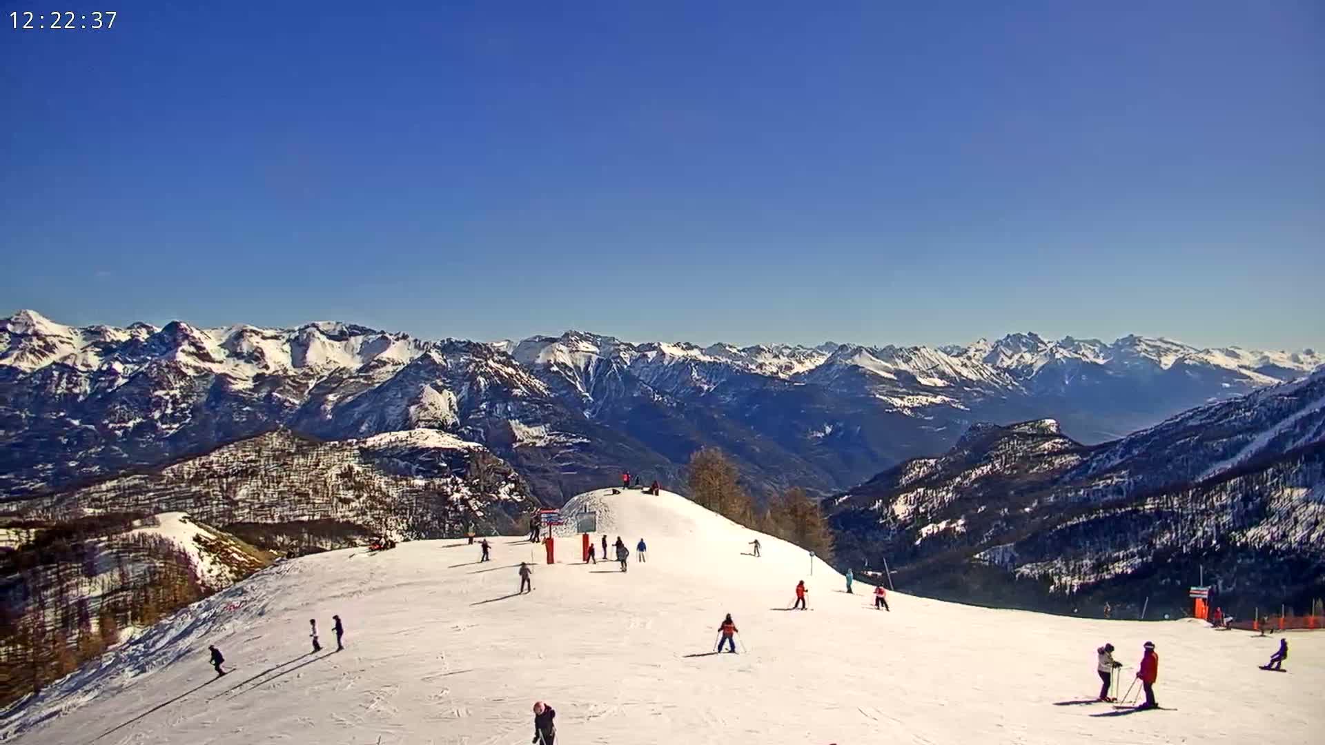 A snow-covered ski slope with several skiers and snowboarders is visible against a backdrop of snow-capped mountains under a clear blue sky.