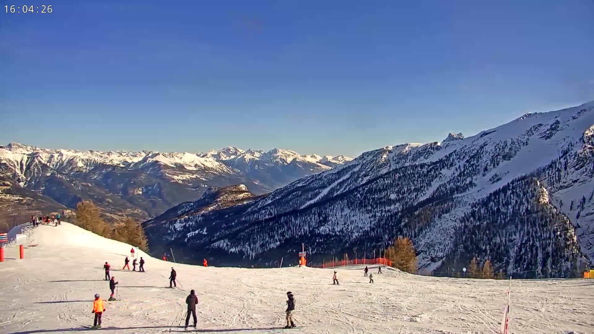 A snow-covered ski slope with skiers and a clear, sunny day, overlooking a mountain range.