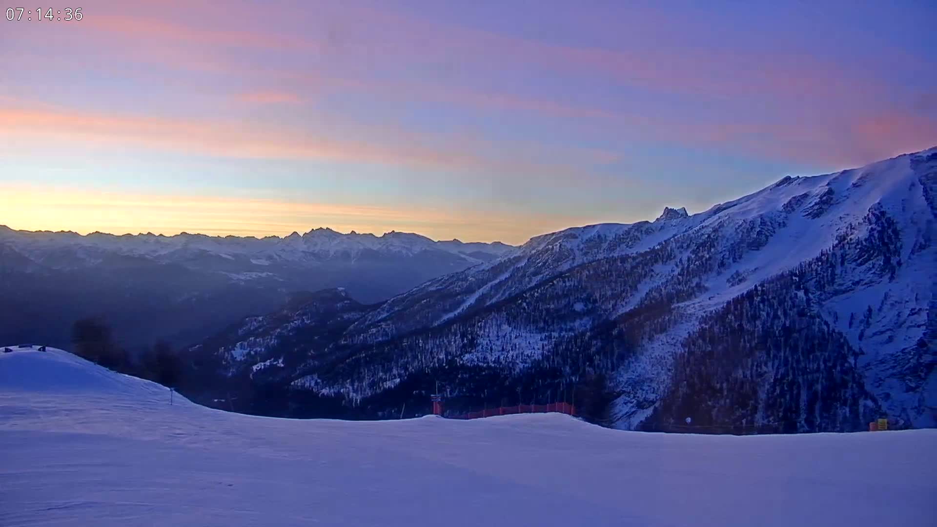A snowy mountain slope overlooks a vast range of snow-covered mountains at sunrise under a purplish-pink sky.