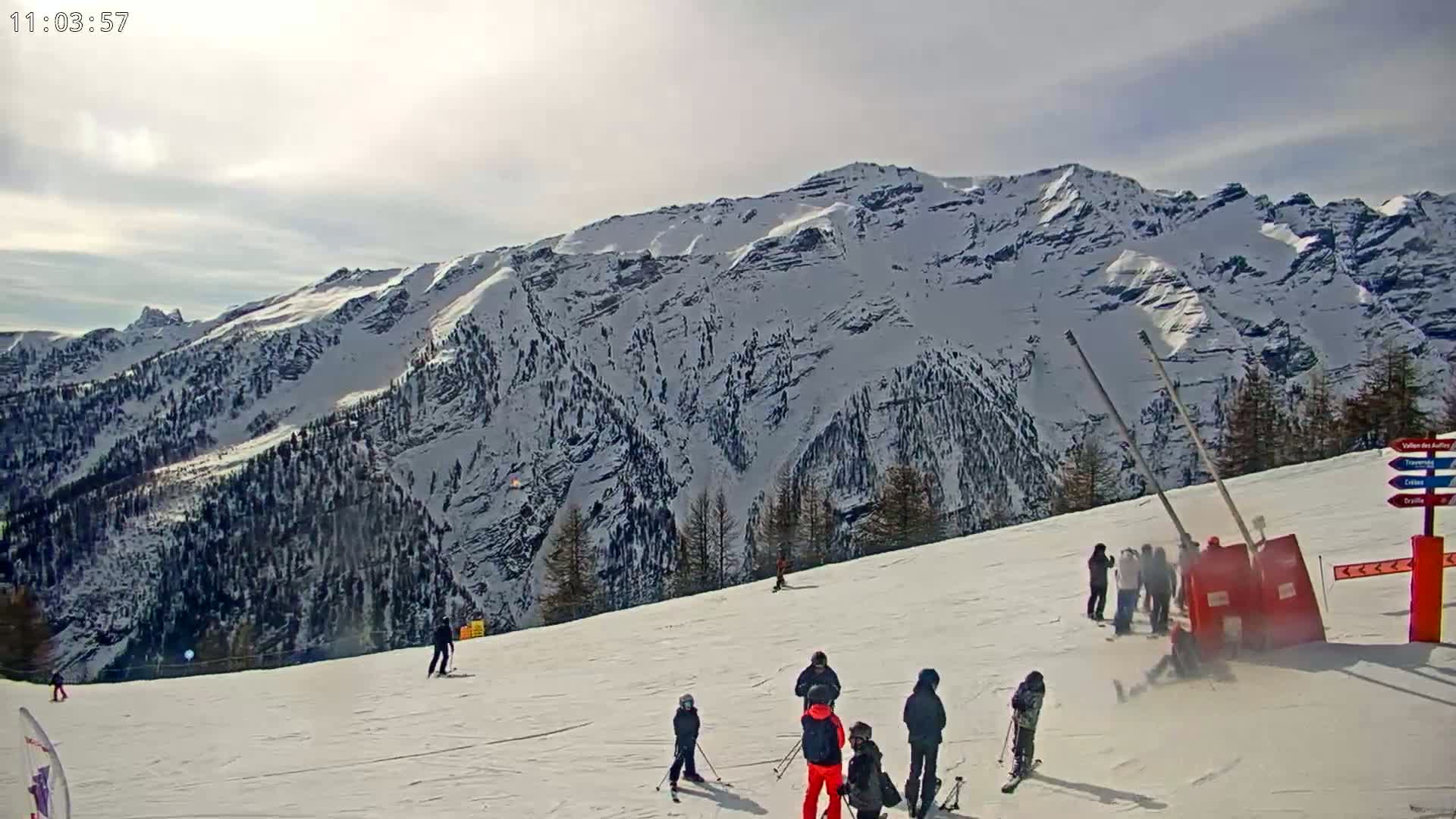 A snowy ski slope with several skiers and snowboarders, a large snow-covered mountain in the background under a mostly sunny sky.