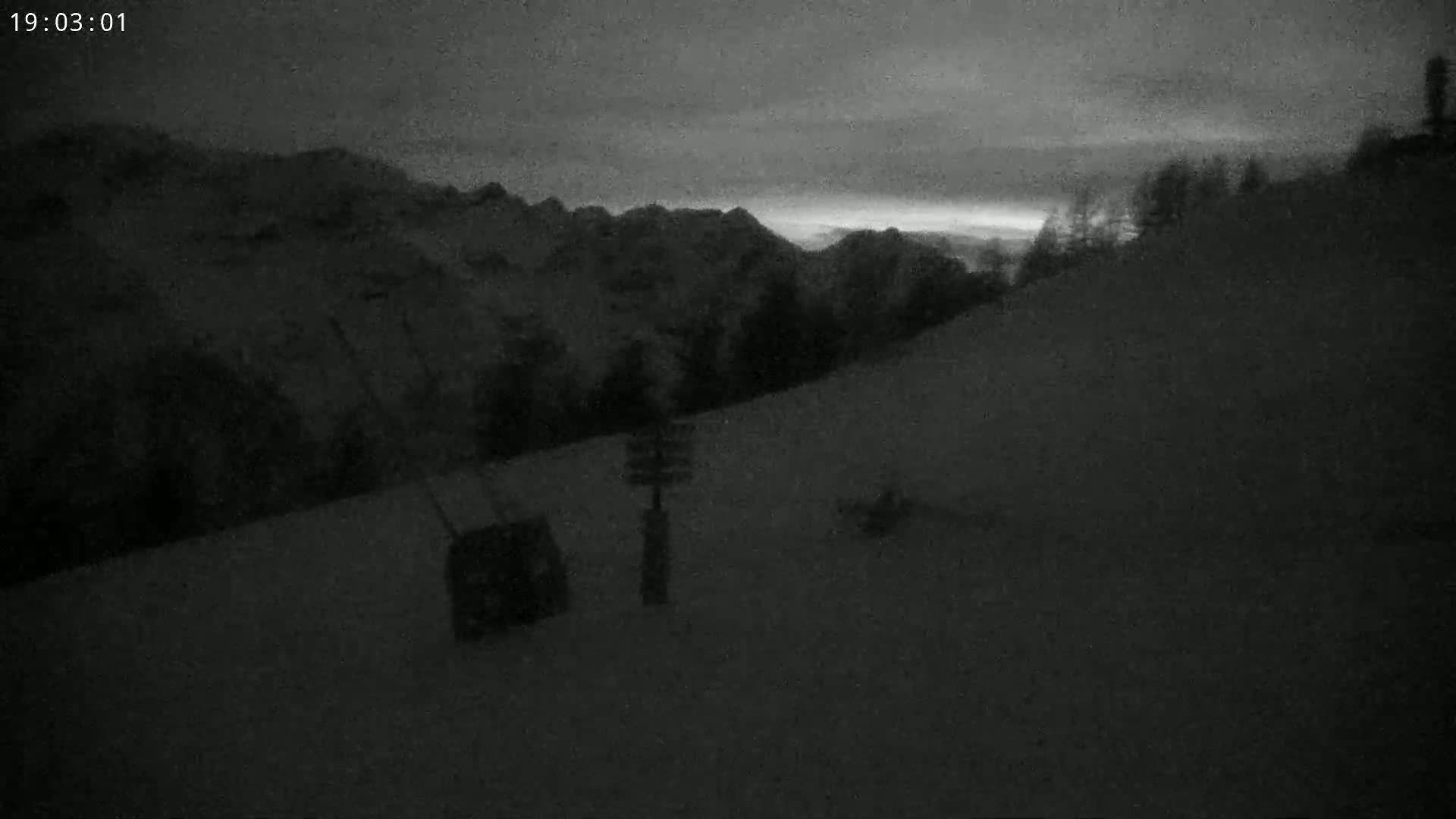 A snow-covered hillside at dusk, with dark mountains in the background and a few pieces of equipment in the foreground.