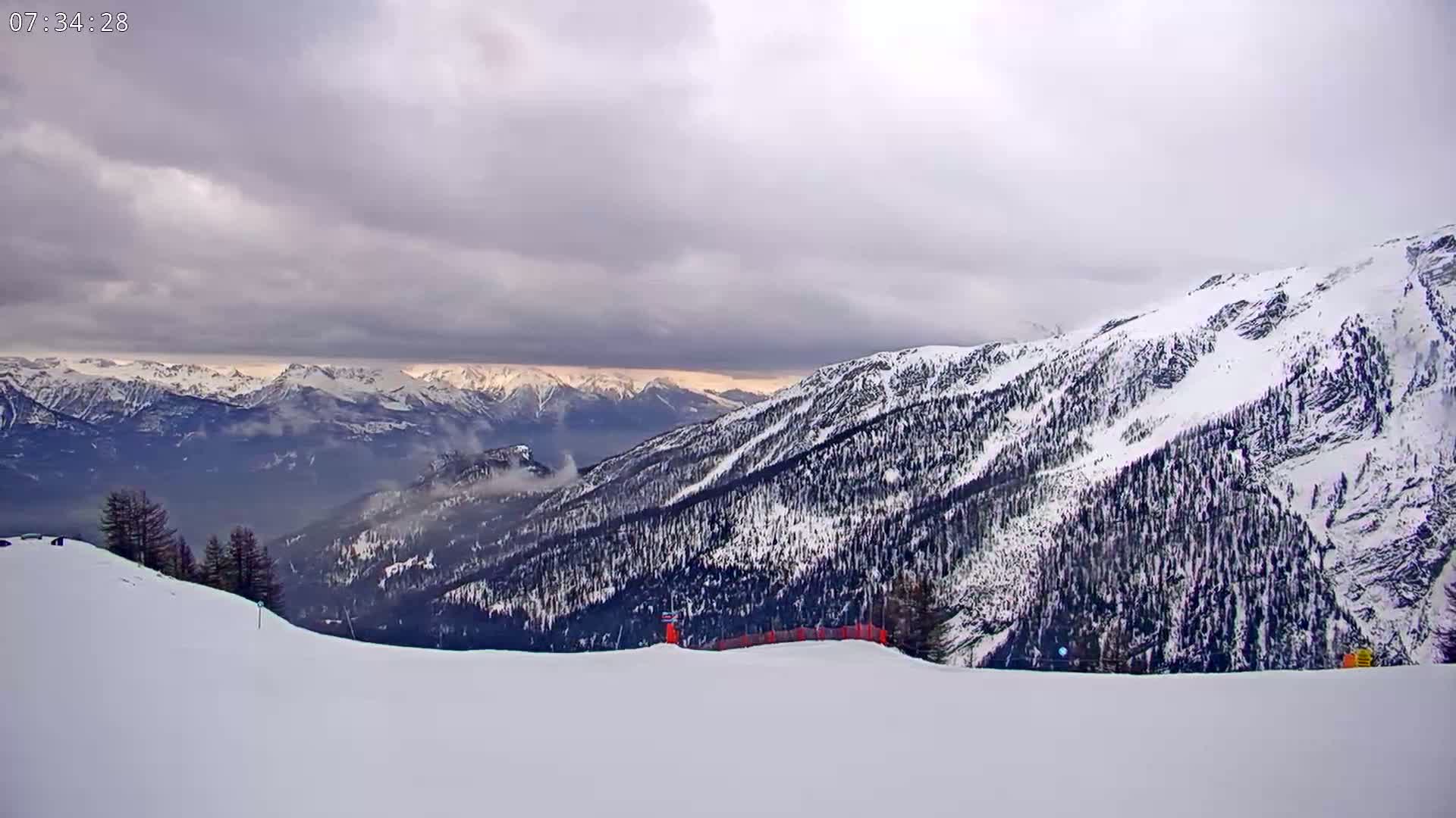 A snow-covered mountain slope overlooks a valley of snow-capped mountains under a cloudy sky.