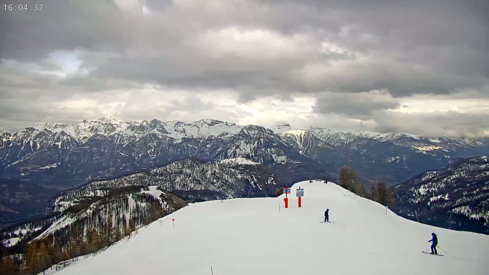 Two skiers are on a snowy mountain slope overlooking a vast, snow-capped mountain range under a cloudy sky.
