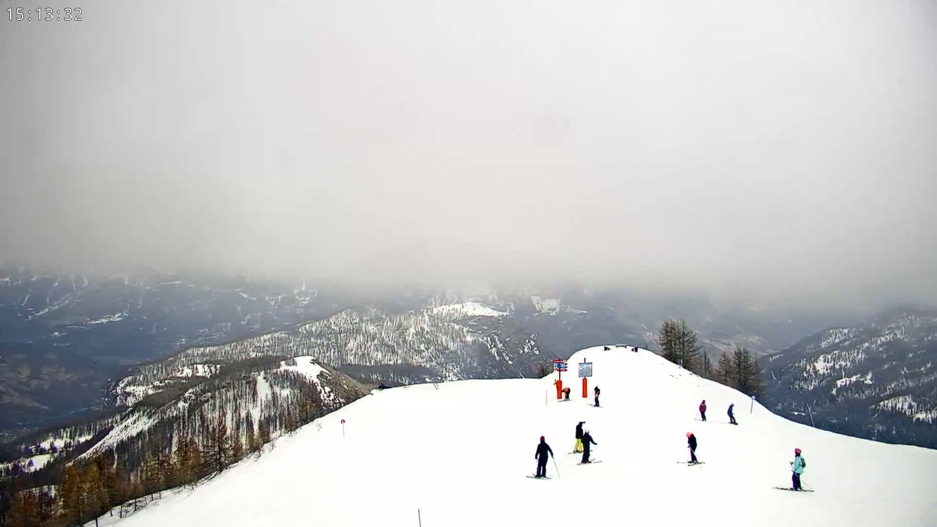 A snowy mountaintop ski slope with several skiers visible on a foggy day.