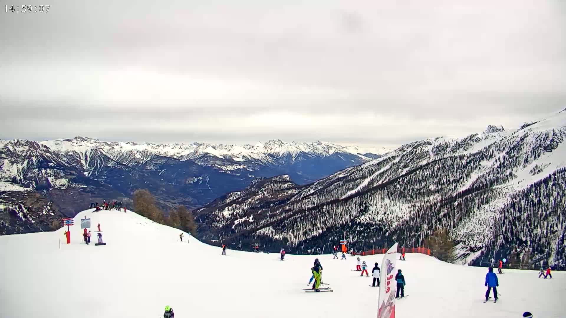 A snowy mountain slope with skiers and snowboarders under an overcast sky, overlooking a vast expanse of snow-covered mountains.