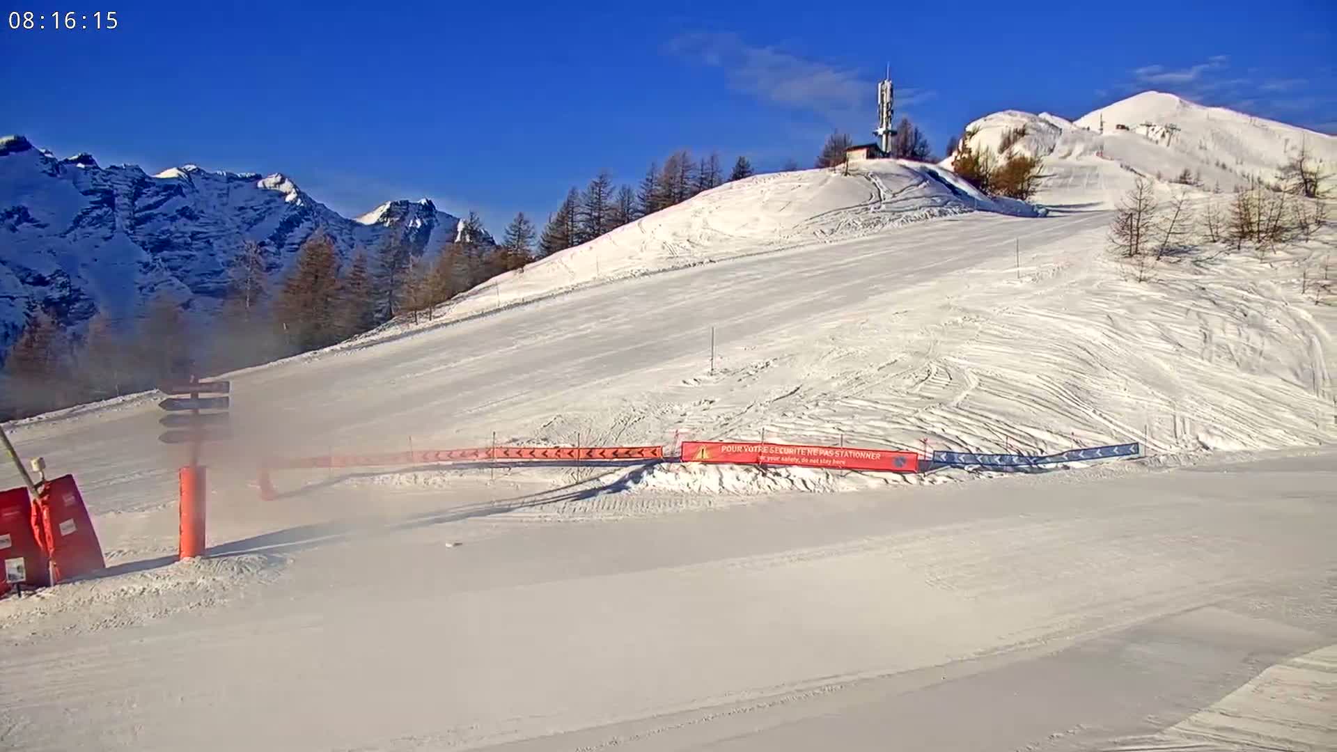 A snow-covered ski slope with a clear blue sky and snow-capped mountains in the background.