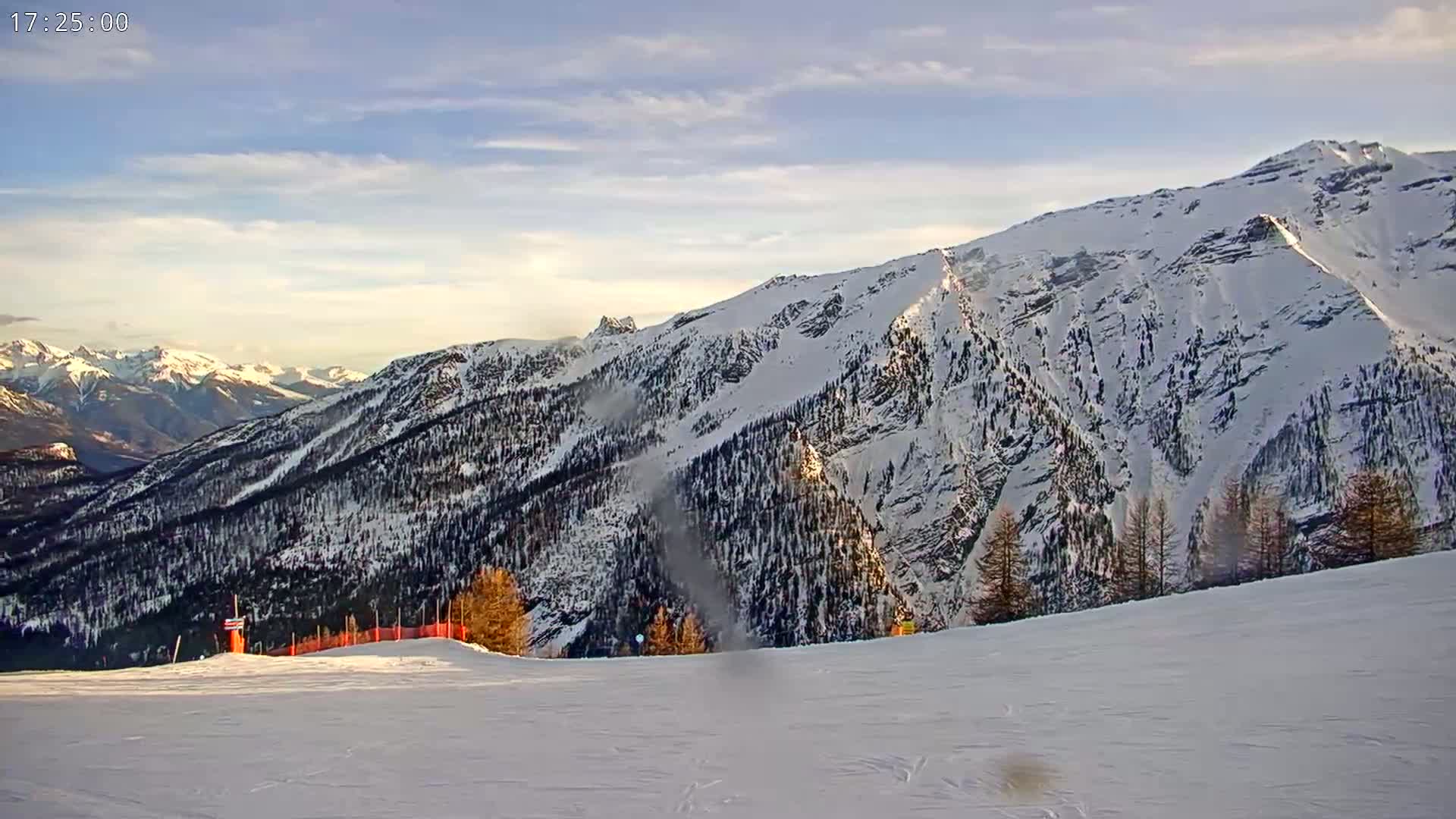 A snow-covered mountain range is visible under a partly cloudy sky, with a flat, snow-covered ski slope in the foreground.