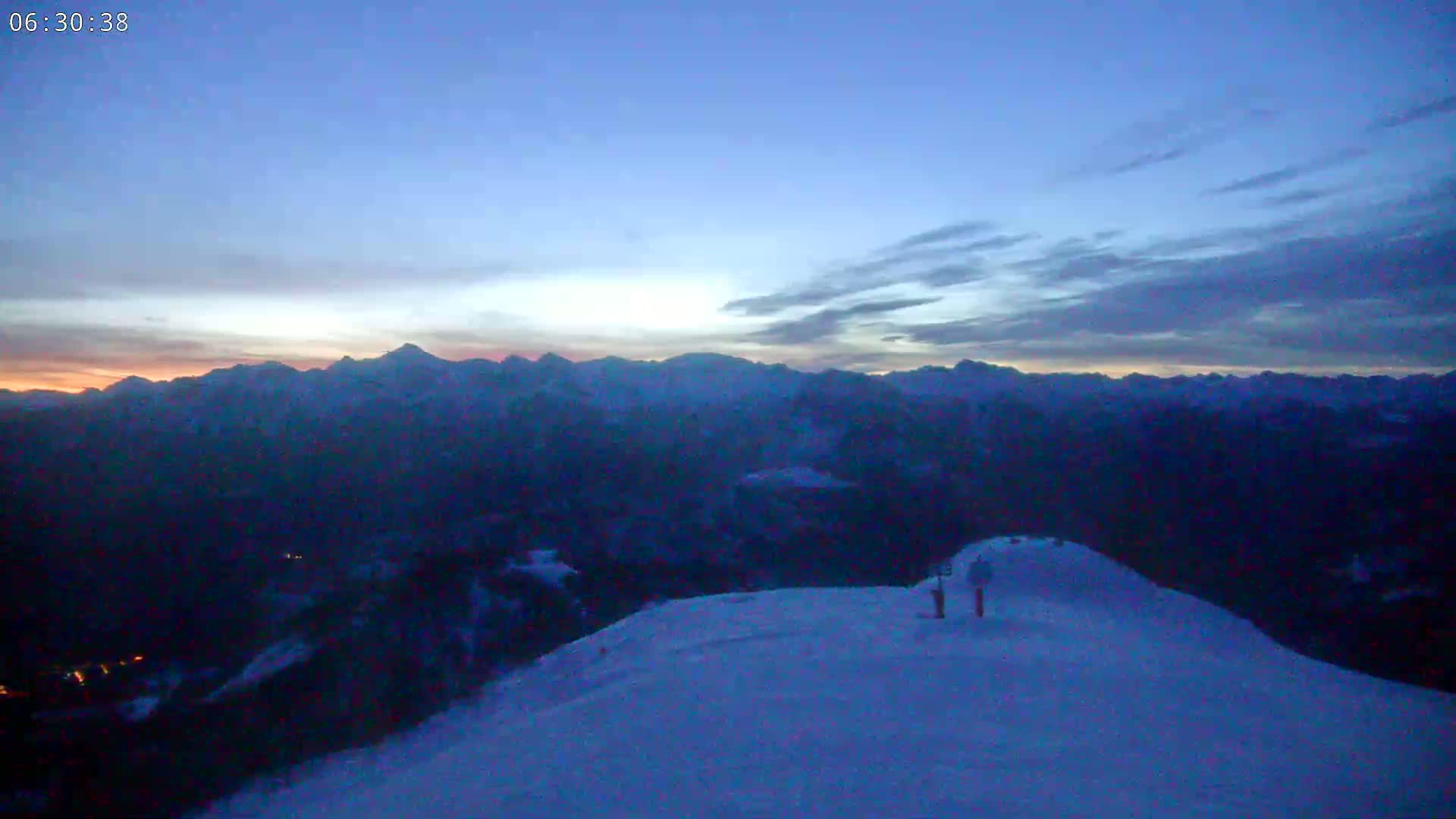 Two figures stand on a snow-covered mountaintop at twilight, overlooking a vast, dark mountain range under a mostly clear, dusky sky.
