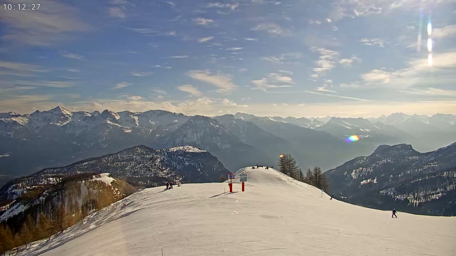 A snowy mountaintop ski slope overlooks a vast expanse of snow-covered mountains under a mostly sunny sky with scattered clouds.