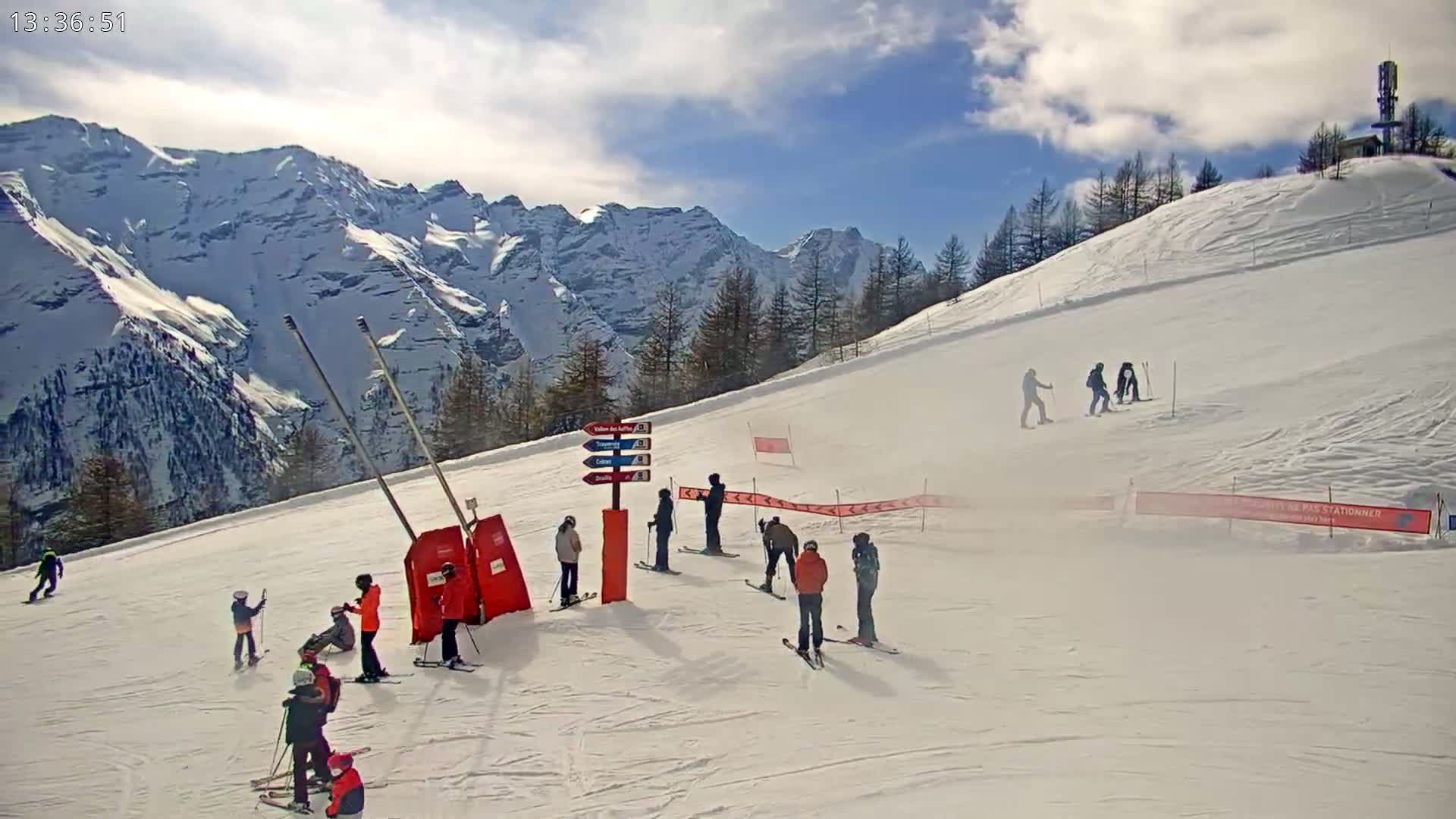A snowy ski slope with several skiers and snowboarders, under a partly cloudy sky, features a backdrop of snow-capped mountains.