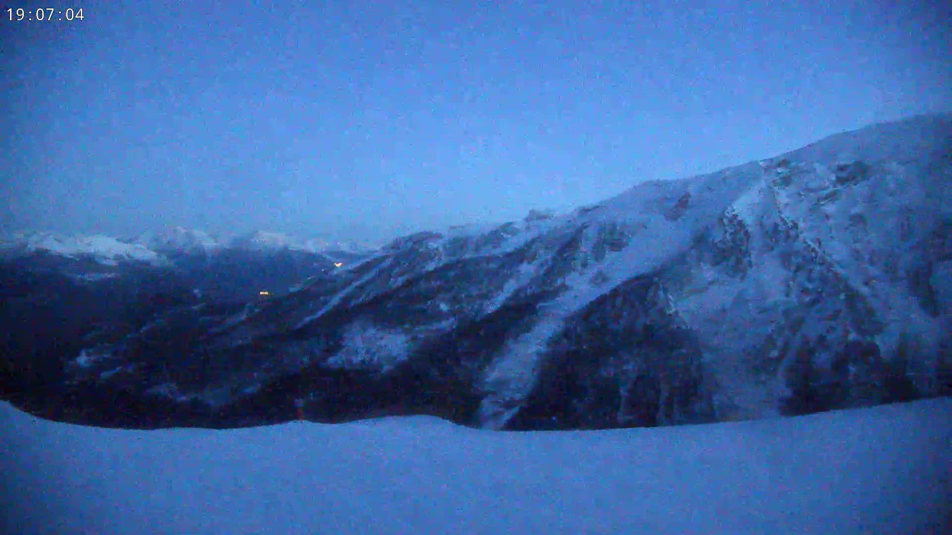 A snow-covered mountain range is visible at twilight, with a few distant lights illuminating a valley below.