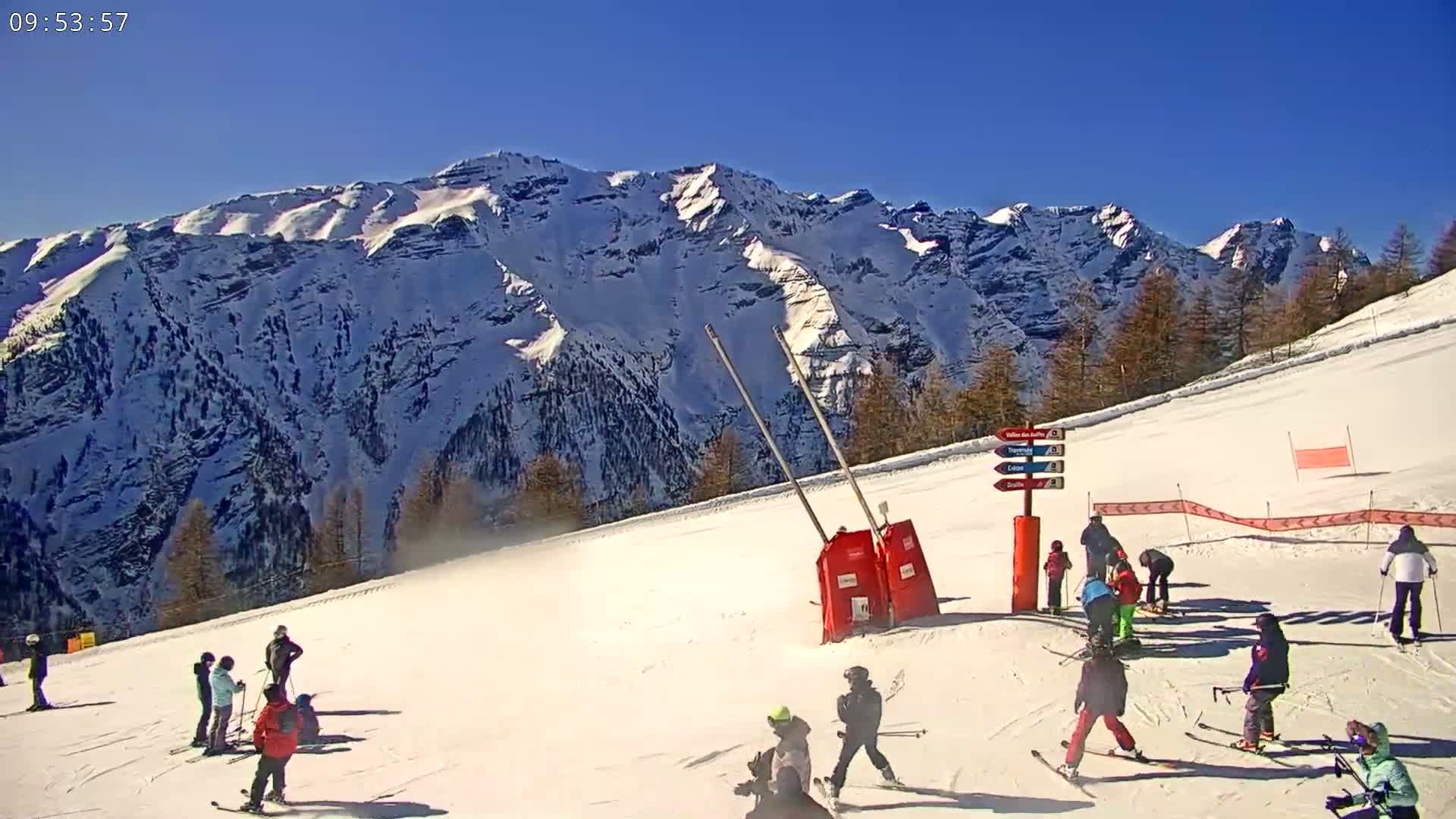 A sunny snow-covered ski slope with skiers and snowboarders, situated in front of a large, snow-capped mountain range.