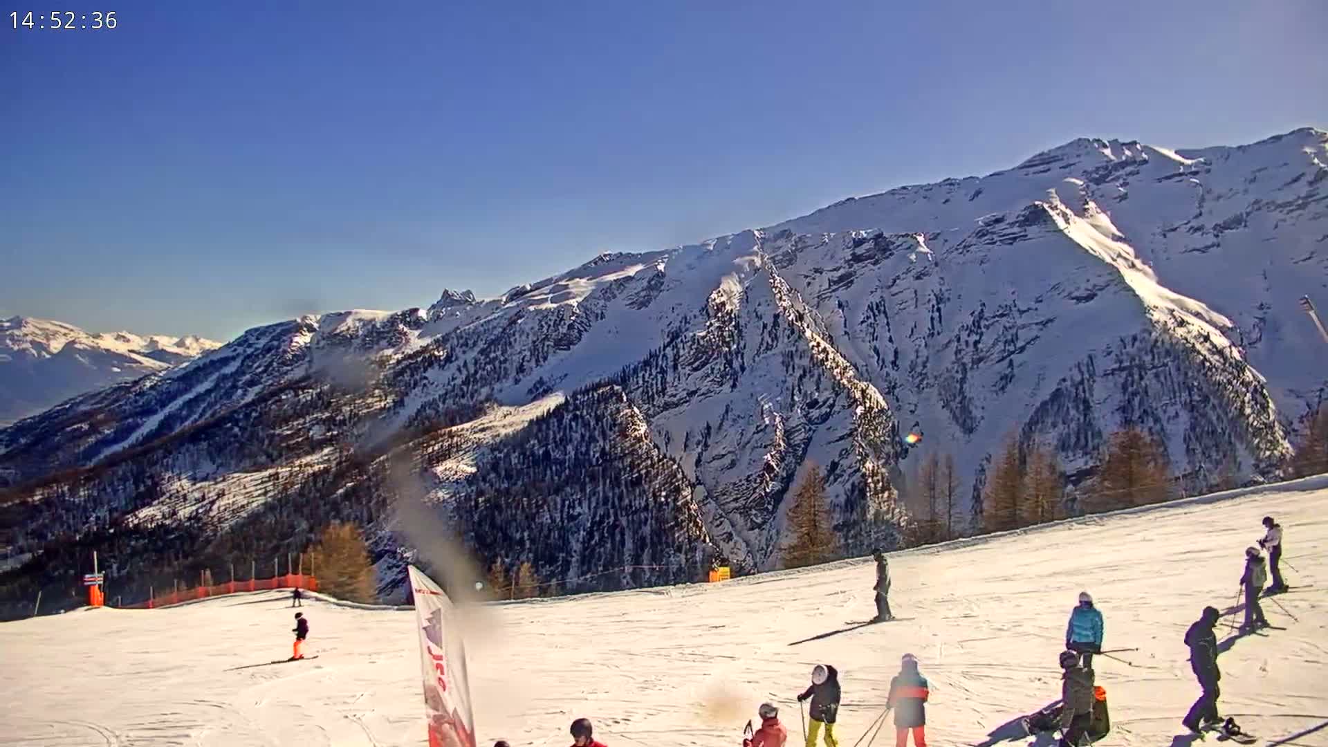 A sunny day on a snow-covered ski slope with skiers and snowboarders, a mountain range in the background.