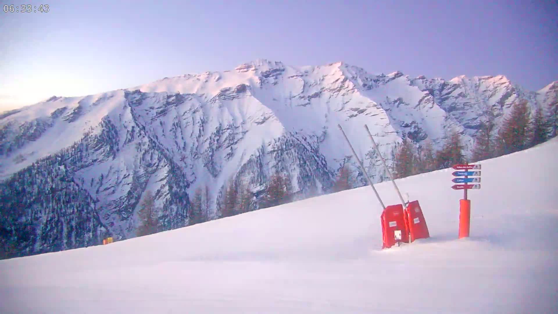 A snow-covered ski slope sits before a large, snow-capped mountain range under a clear, pale blue sky.