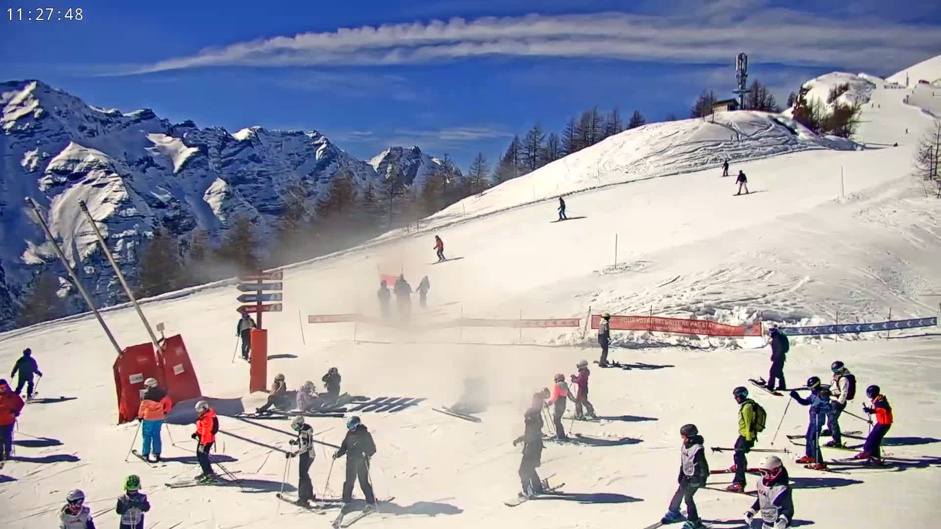 A sunny day on a snow-covered ski slope shows numerous skiers and snowboarders, many in groups, against a backdrop of snow-capped mountains.