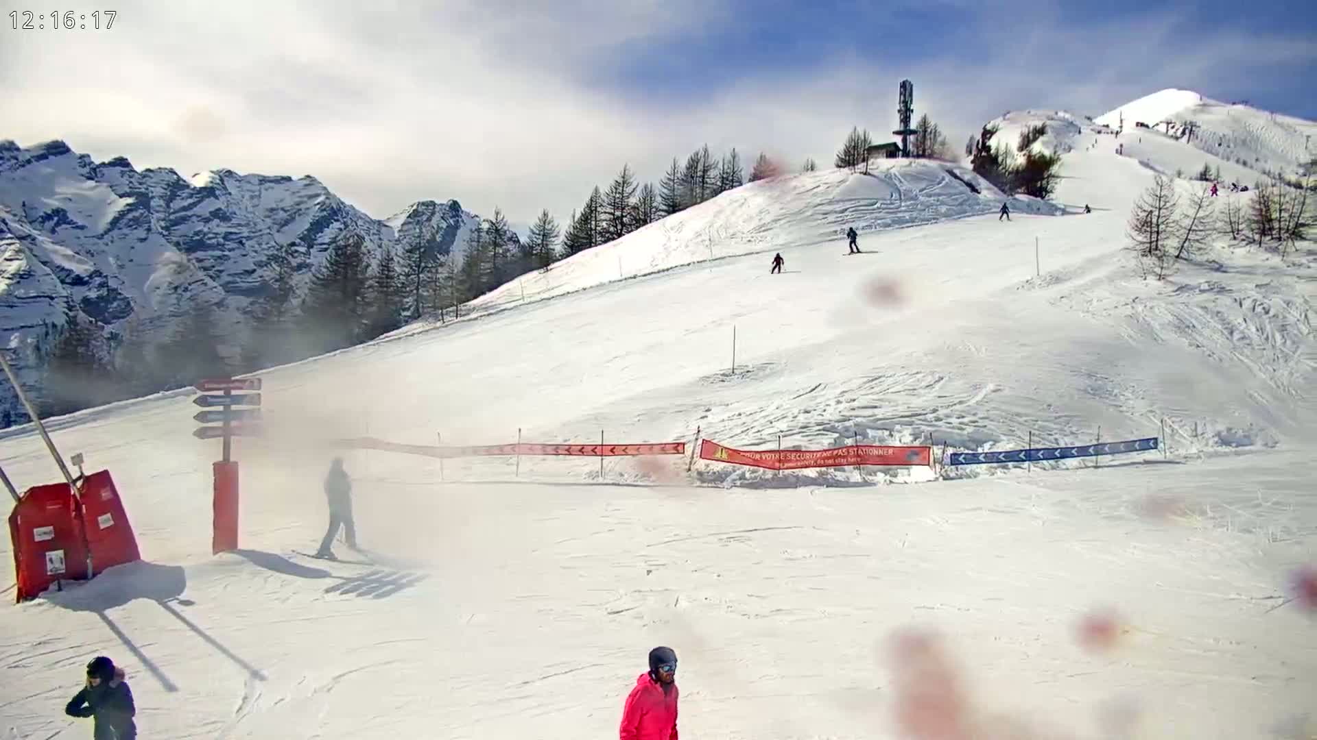 A snowy ski slope with several skiers visible against a backdrop of mountainous terrain under a partly cloudy sky.