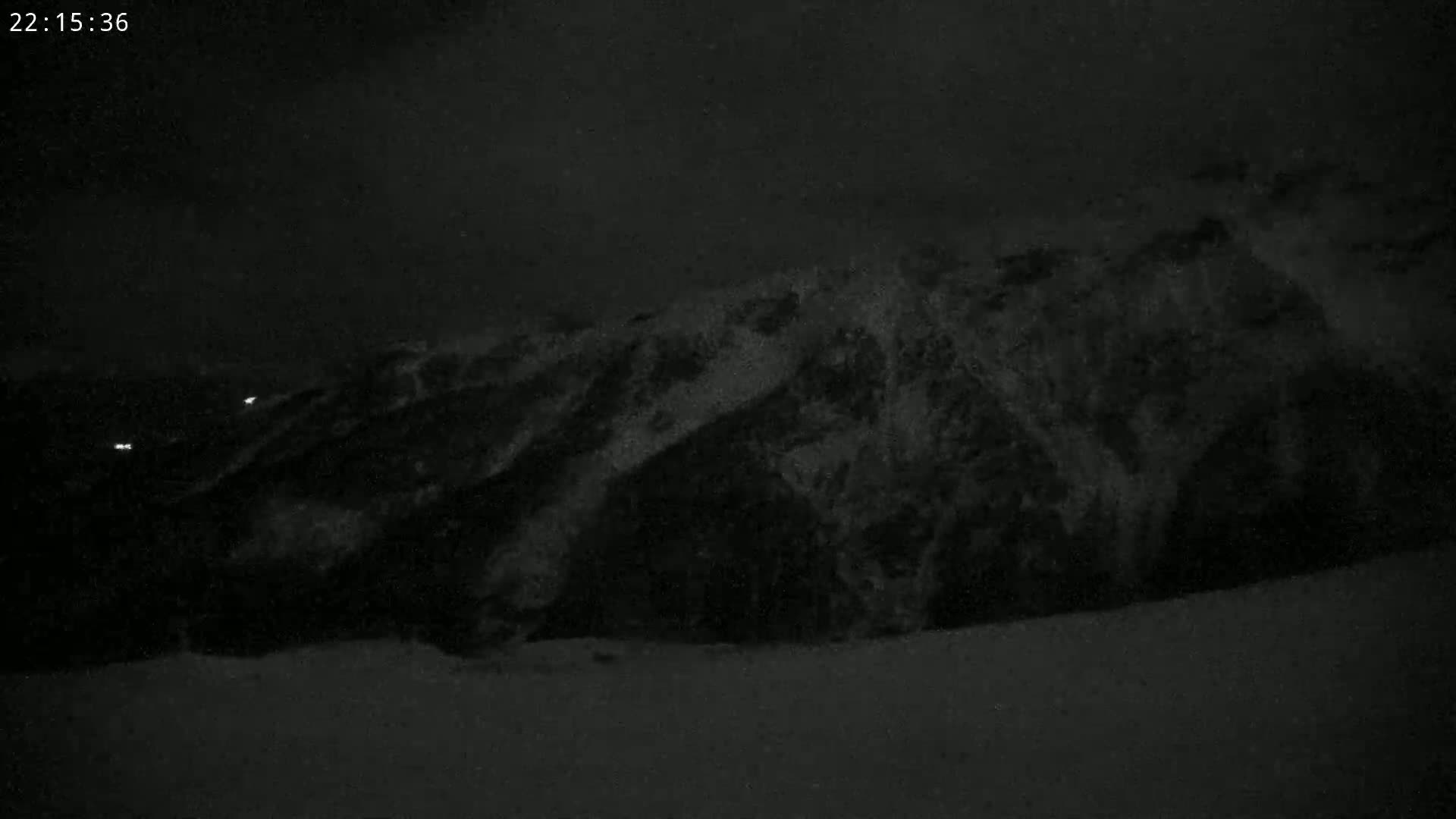 A dark, nighttime view of a snow-covered mountainside with faint lights visible in the distance.