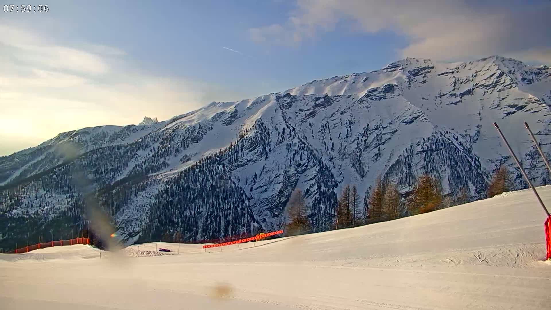 A snow-covered mountain range is visible under a mostly clear, bright sky, with a snowy ski slope in the foreground.
