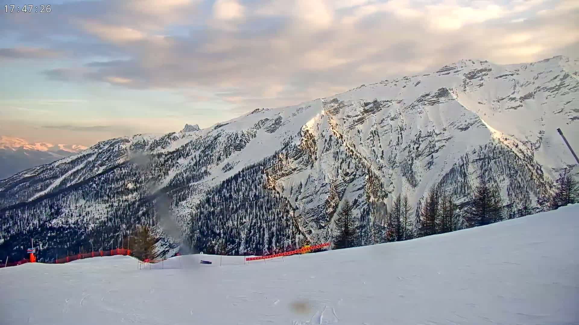 A snow-covered mountain range is visible under a partly cloudy sunset sky, with a groomed ski slope in the foreground.