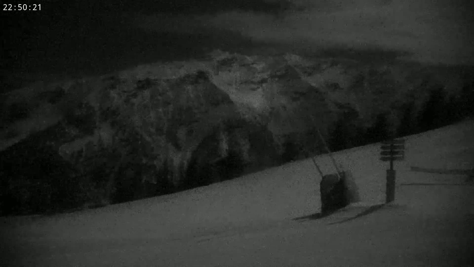 A nighttime, snowy landscape shows a ski lift and a directional signpost in the foreground, with dark mountains in the background under a cloudy sky.