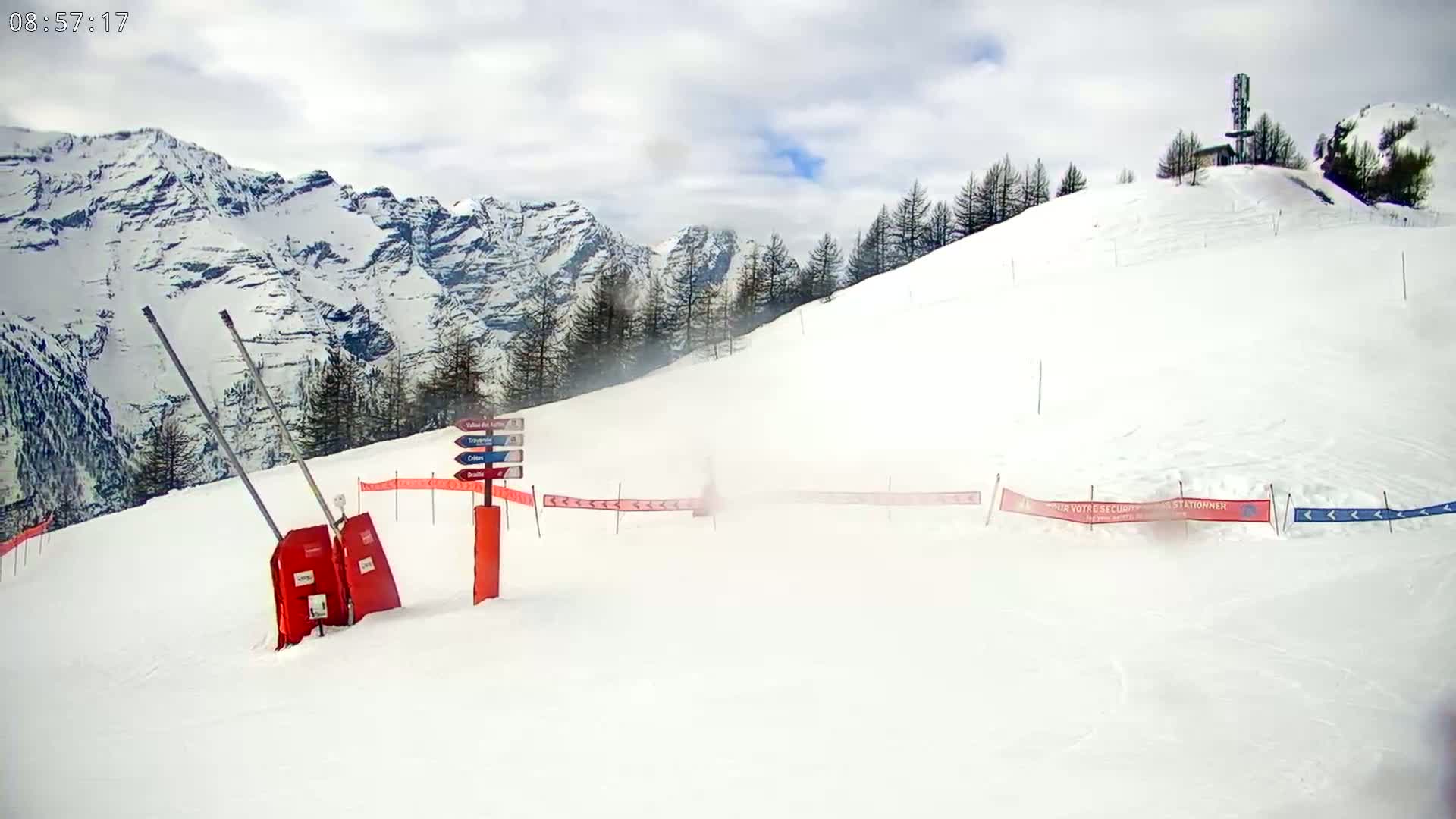 A snow-covered ski slope with a mountain range in the background under partly cloudy skies.