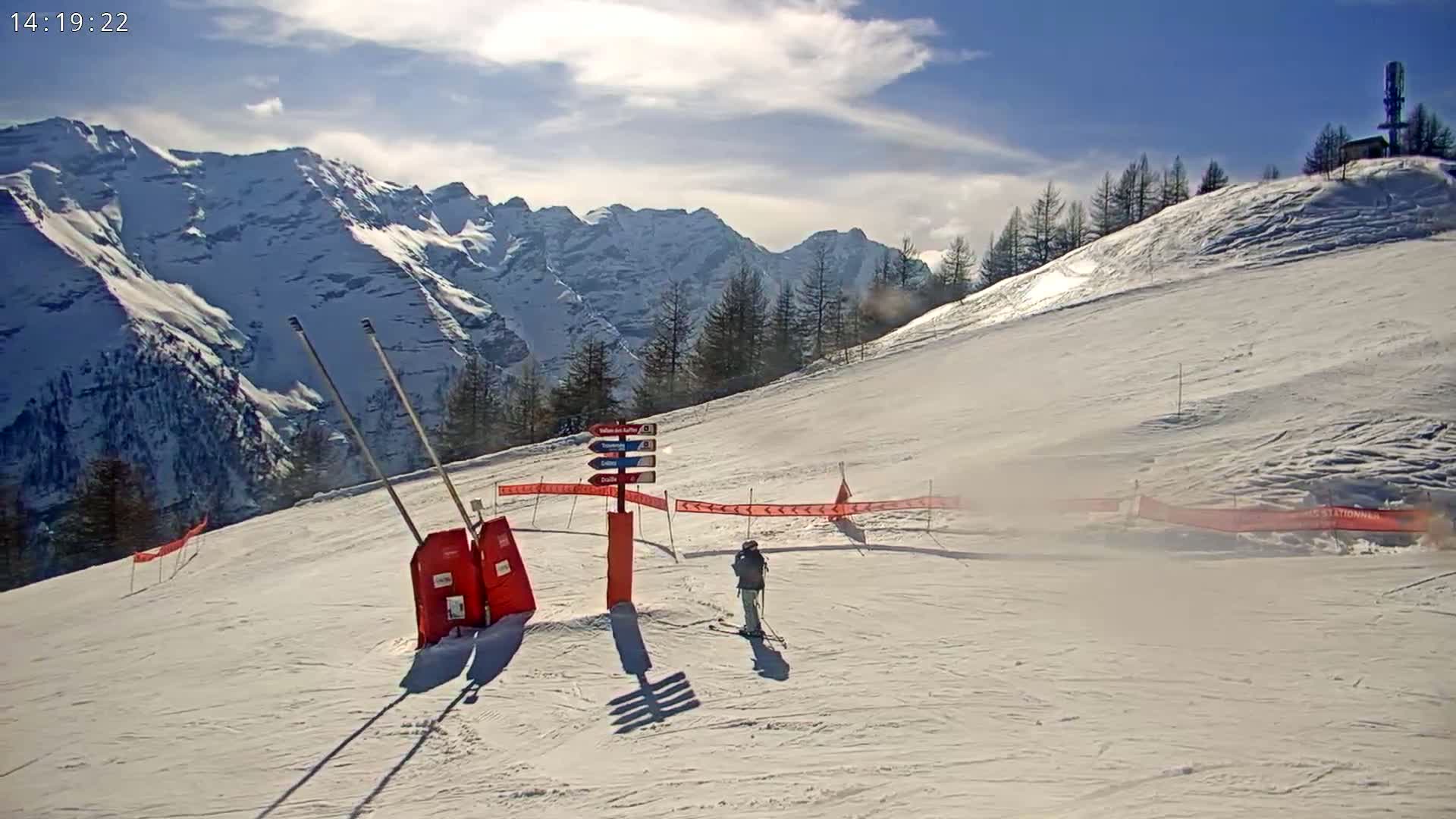 A skier stands on a snow-covered ski slope with mountains in the background under a mostly sunny sky.
