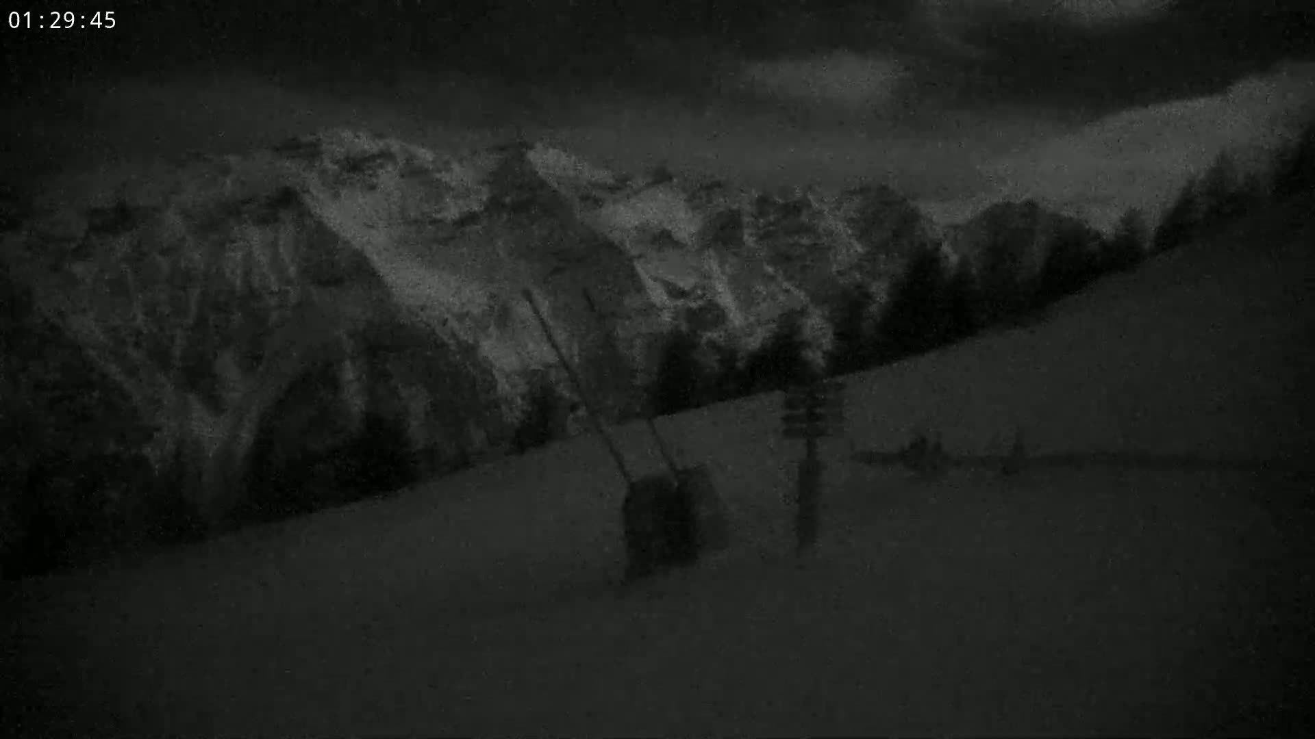 A nighttime, snow-covered hillside slopes down towards a dark, snow-covered mountain range under a cloudy sky.