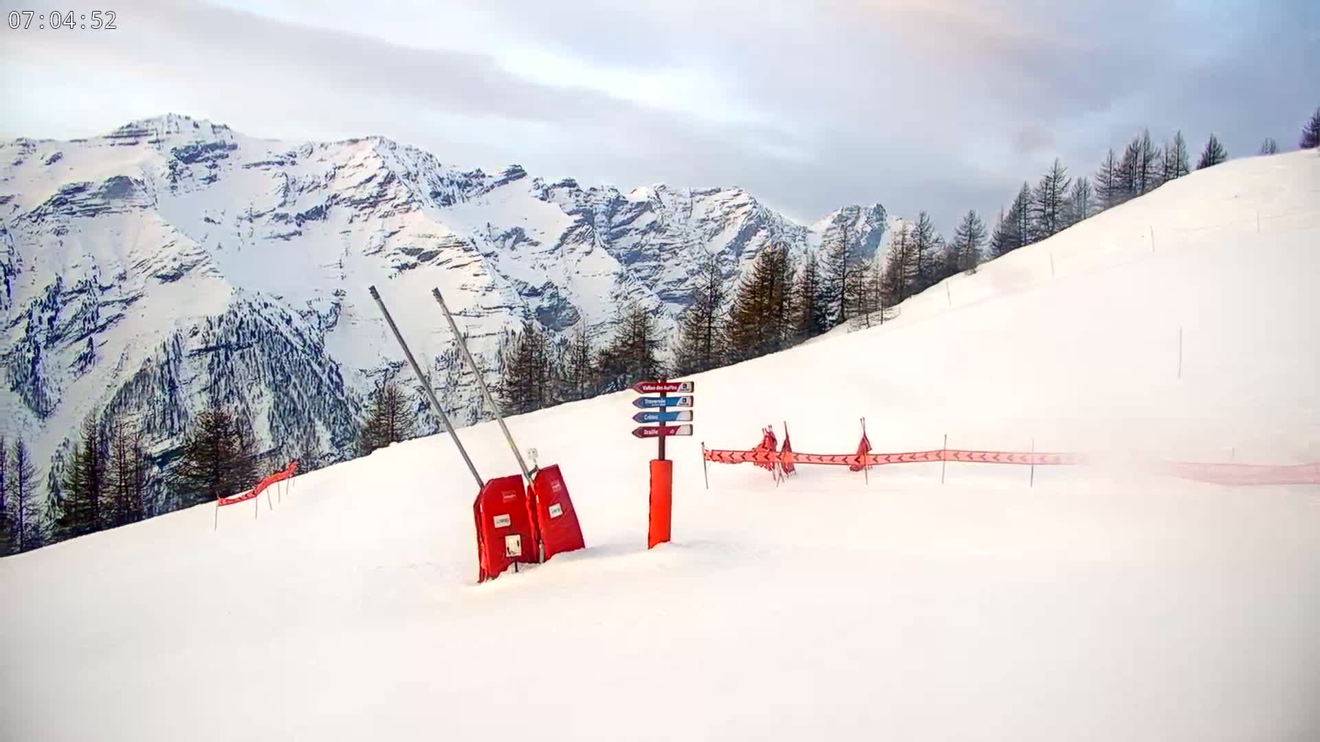A snow-covered ski slope with a signpost and red safety gates is situated in front of a mountainous landscape under a mostly cloudy sky.