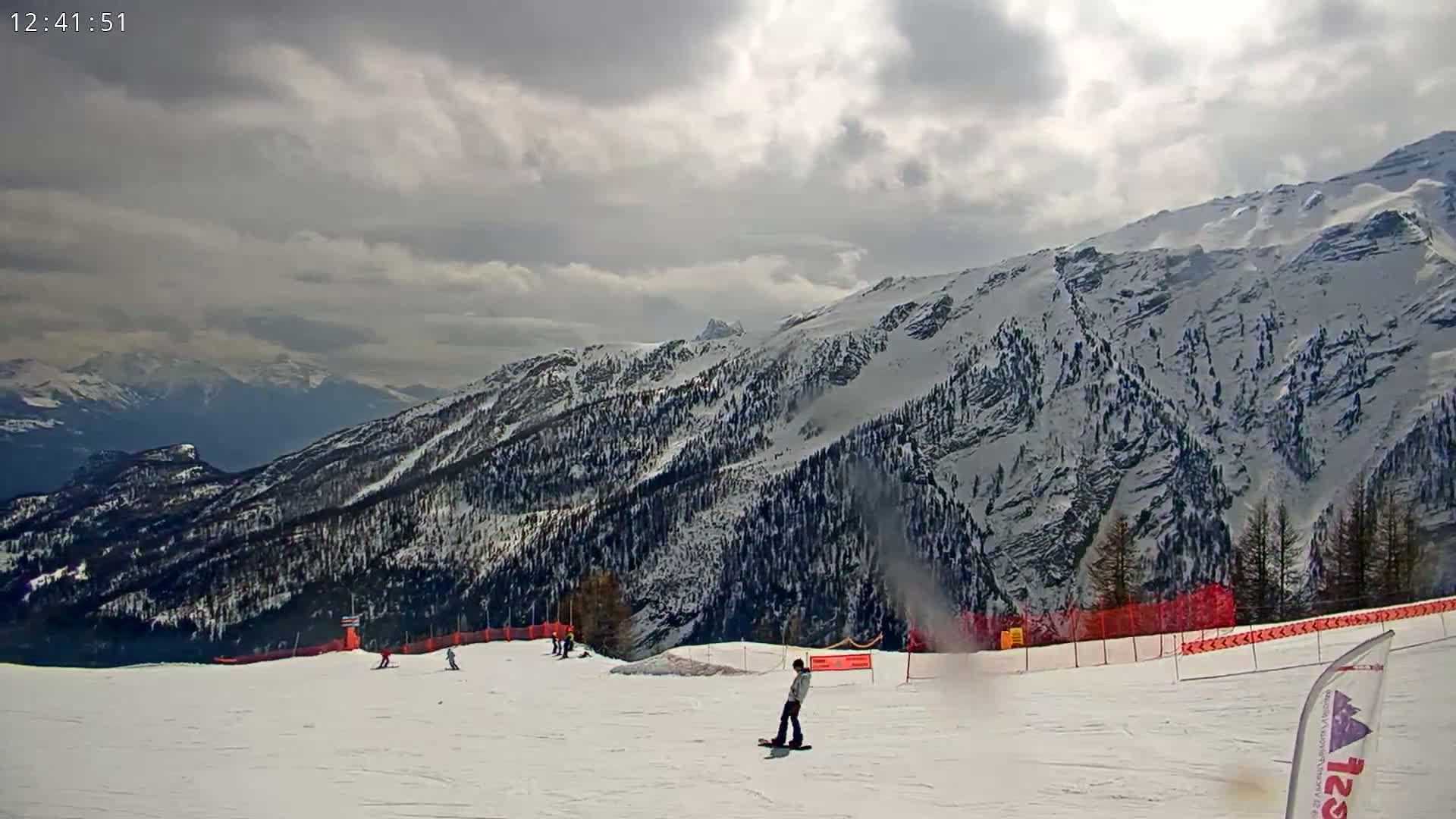 A snowboarder stands on a snowy ski slope overlooking snow-covered mountains under a cloudy sky.