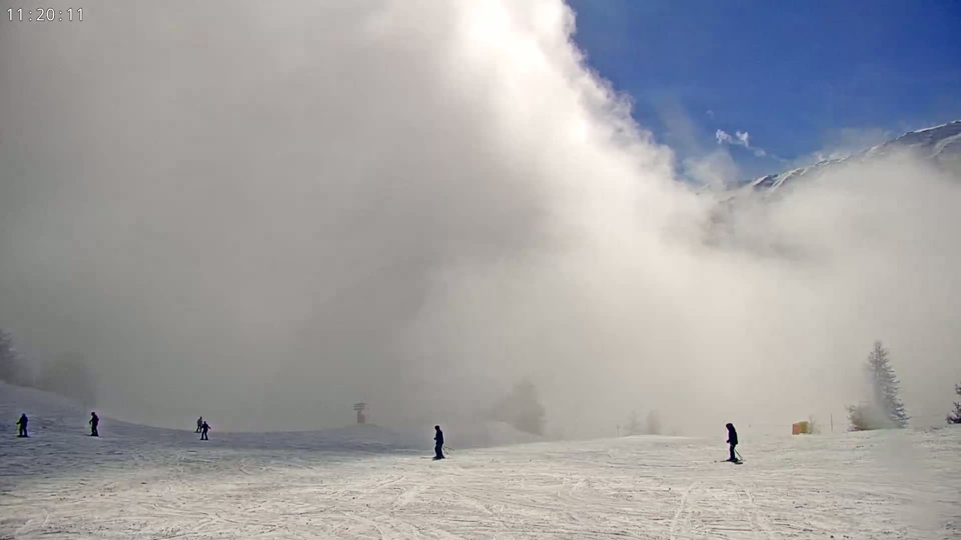 A snowy ski slope is partially obscured by heavy fog under a partly sunny sky, with several skiers visible on the slope.