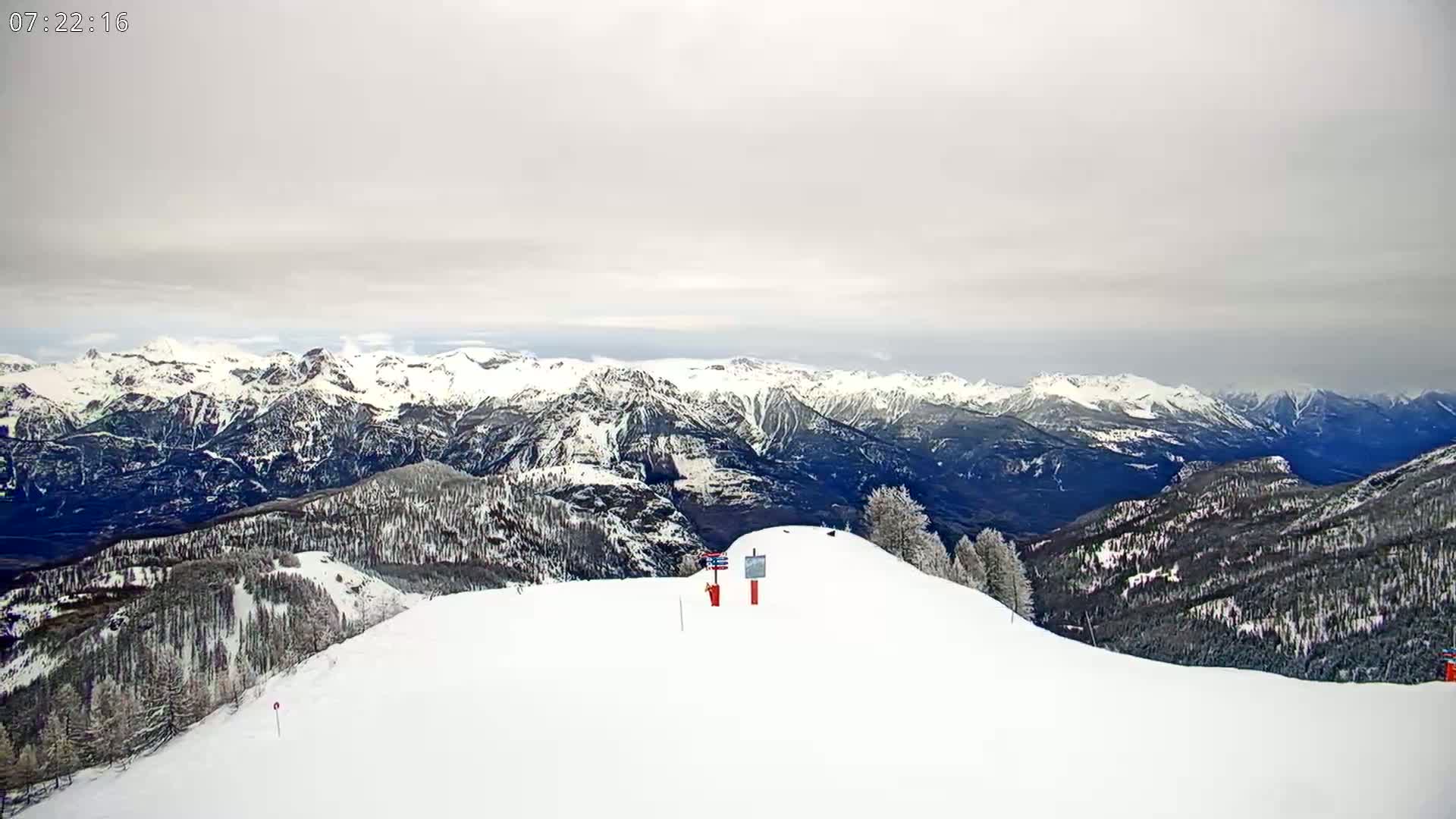 A snow-covered mountaintop with a ski slope overlooks a vast range of snow-capped mountains under an overcast sky.