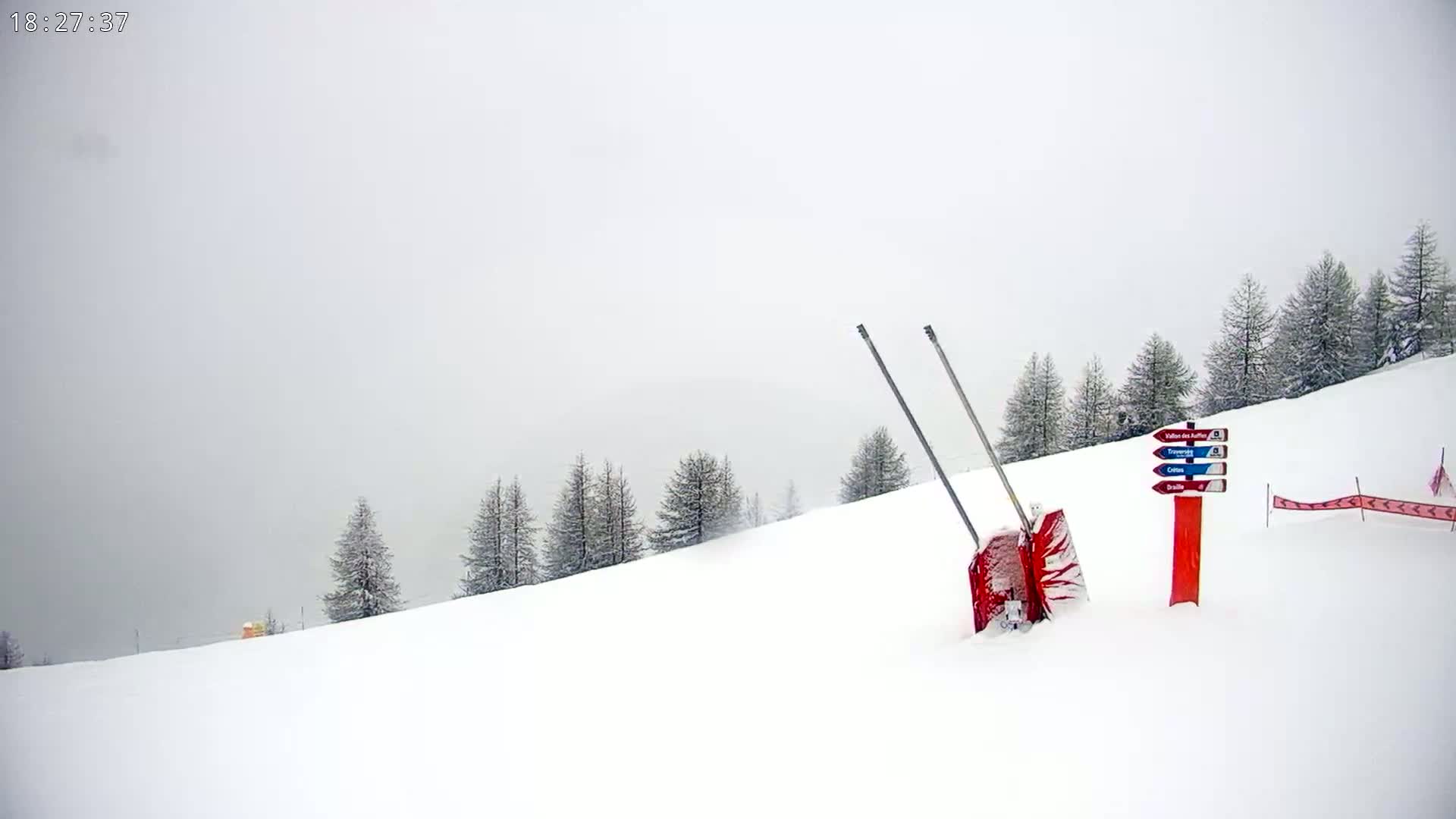 A snow-covered ski slope with a red snow plow partially buried in the snow, a trail marker, and a line of evergreen trees in the background under overcast skies.