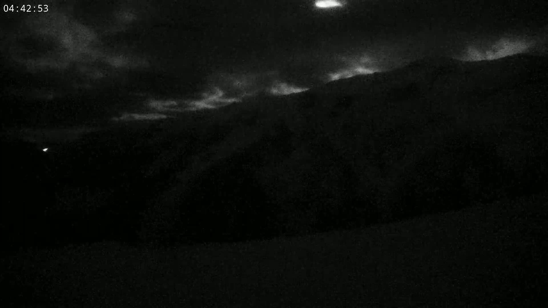 A dark, nighttime landscape shows a mountain silhouetted against a cloudy sky.