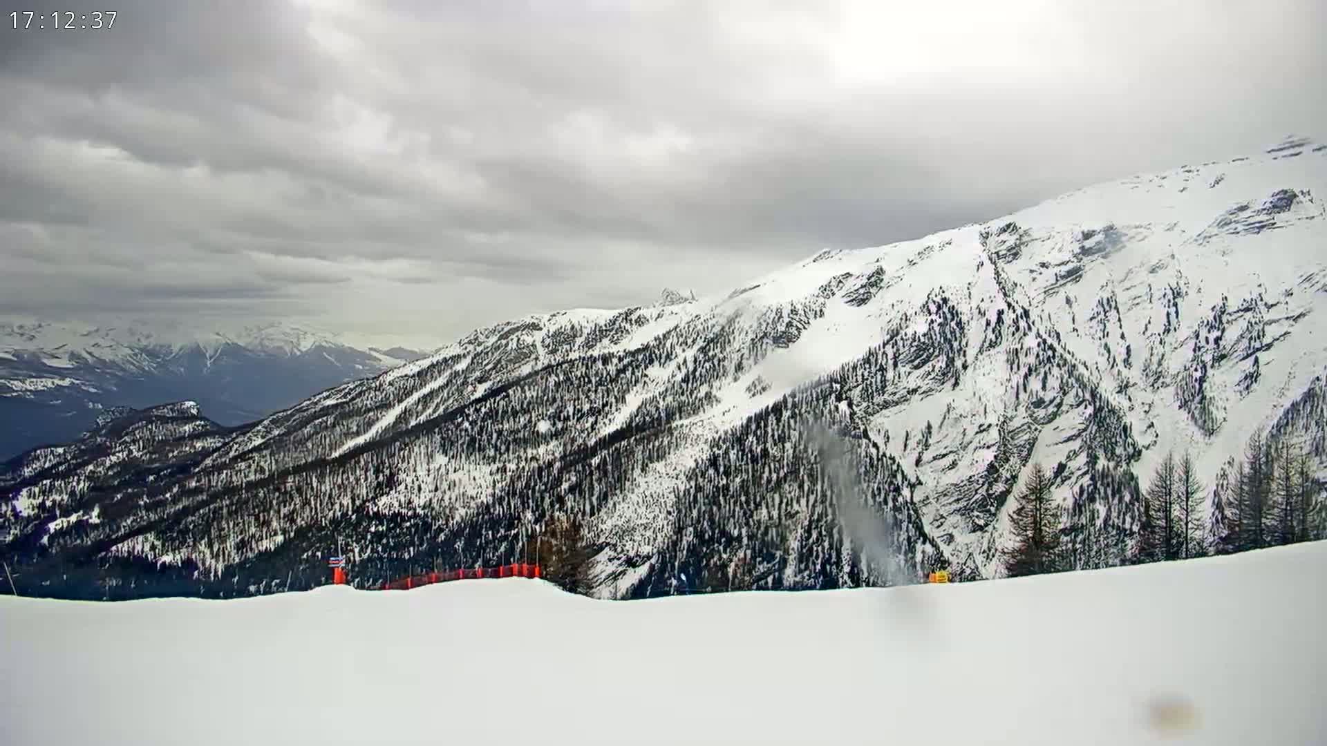 A snow-covered mountain range under a cloudy sky, with a section of a ski slope in the foreground.
