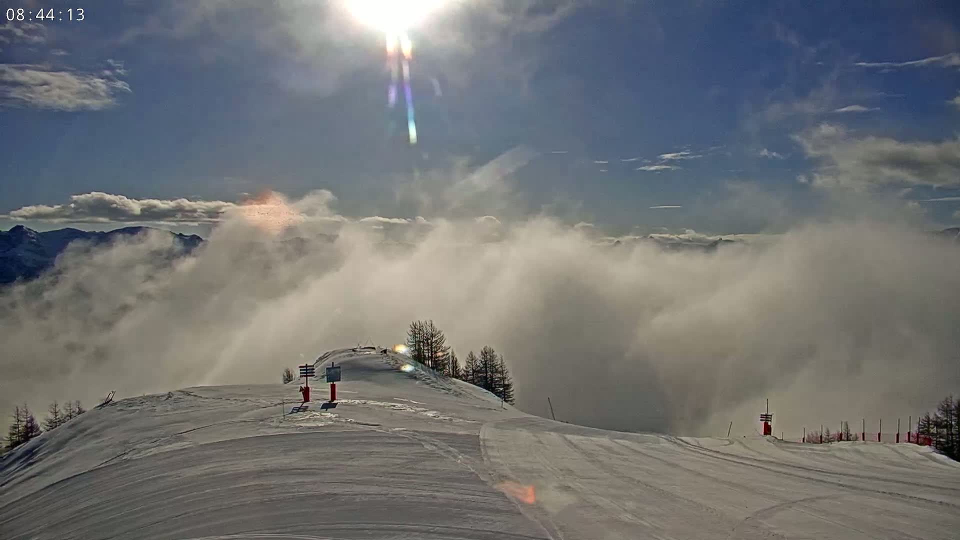 A sunny, partly cloudy day reveals a snow-covered mountain peak with a ski slope, partially obscured by low-lying clouds in the valley below.