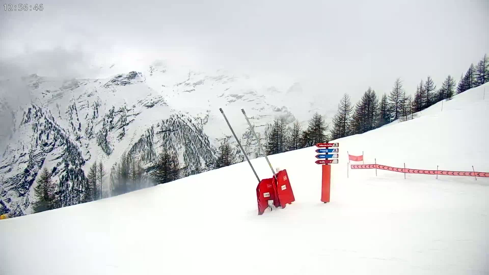 A snowy mountain slope with overcast skies features a pair of red snow removal devices near a trail marker and directional fencing.