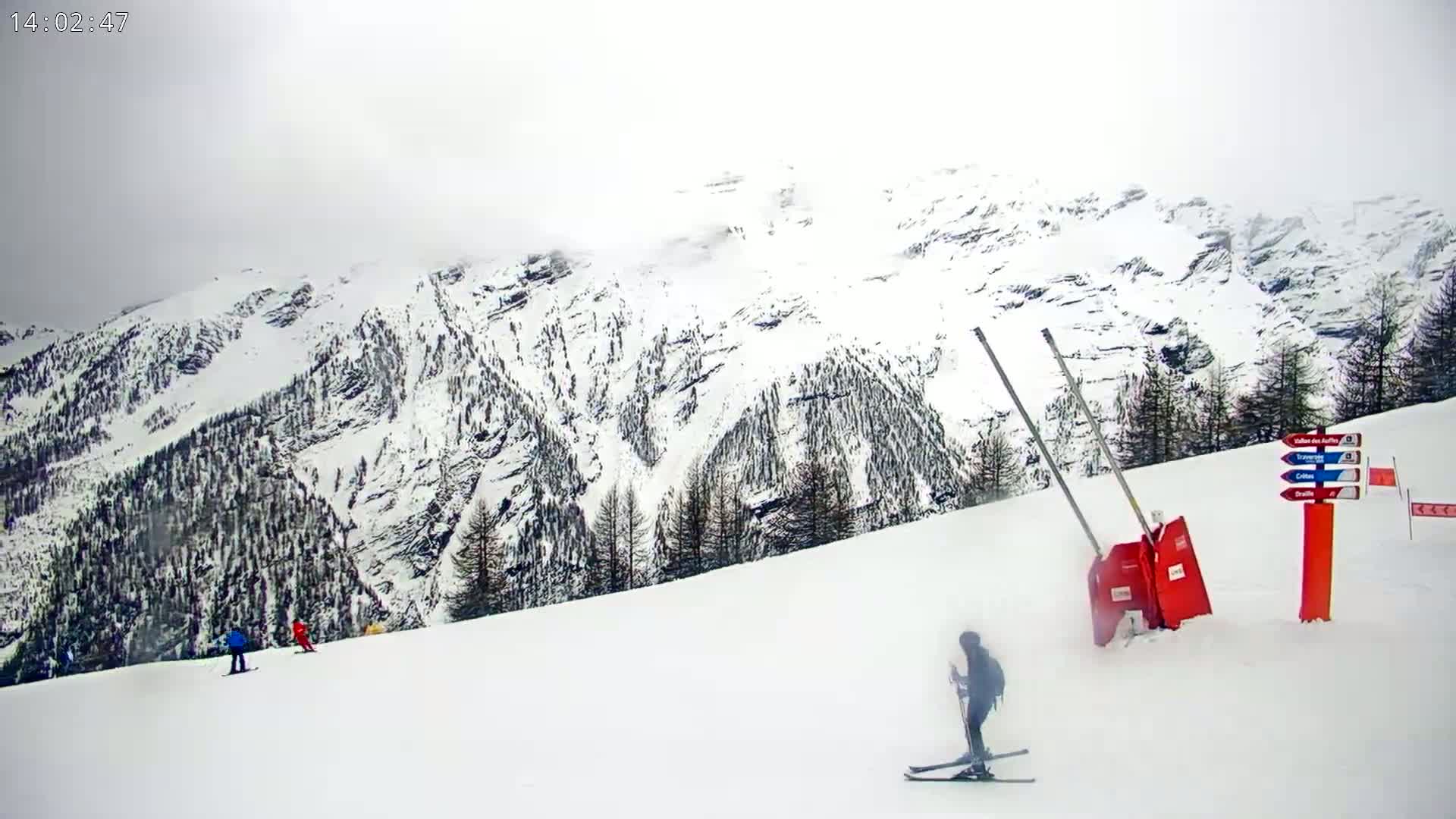 A snowy mountain range is visible in the background, with several skiers on a snow-covered slope in the foreground under overcast skies.