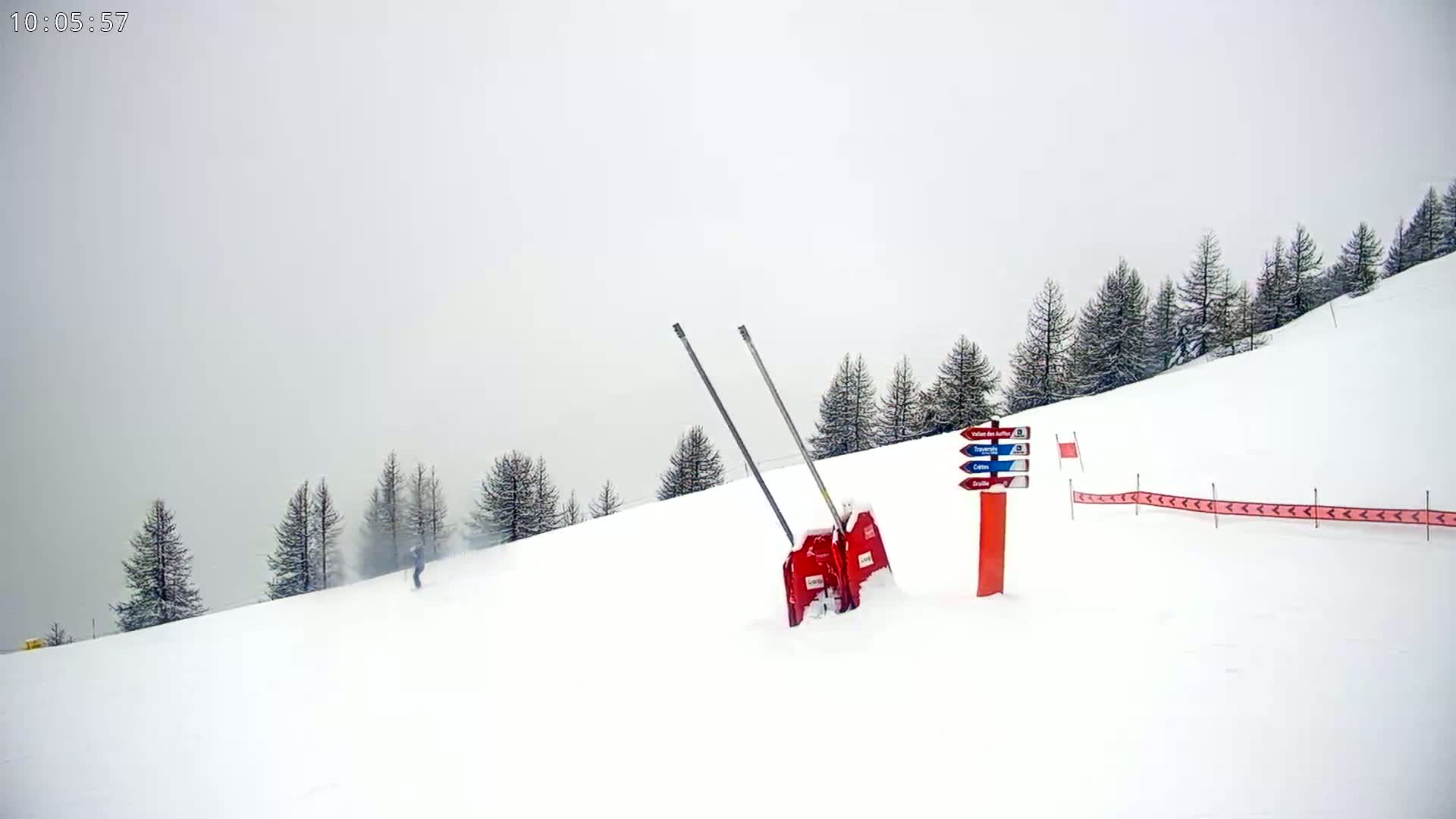 A snowy ski slope with a few skiers, snow-covered trees, a trail marker, and a snow-covered ski lift gate under an overcast sky.